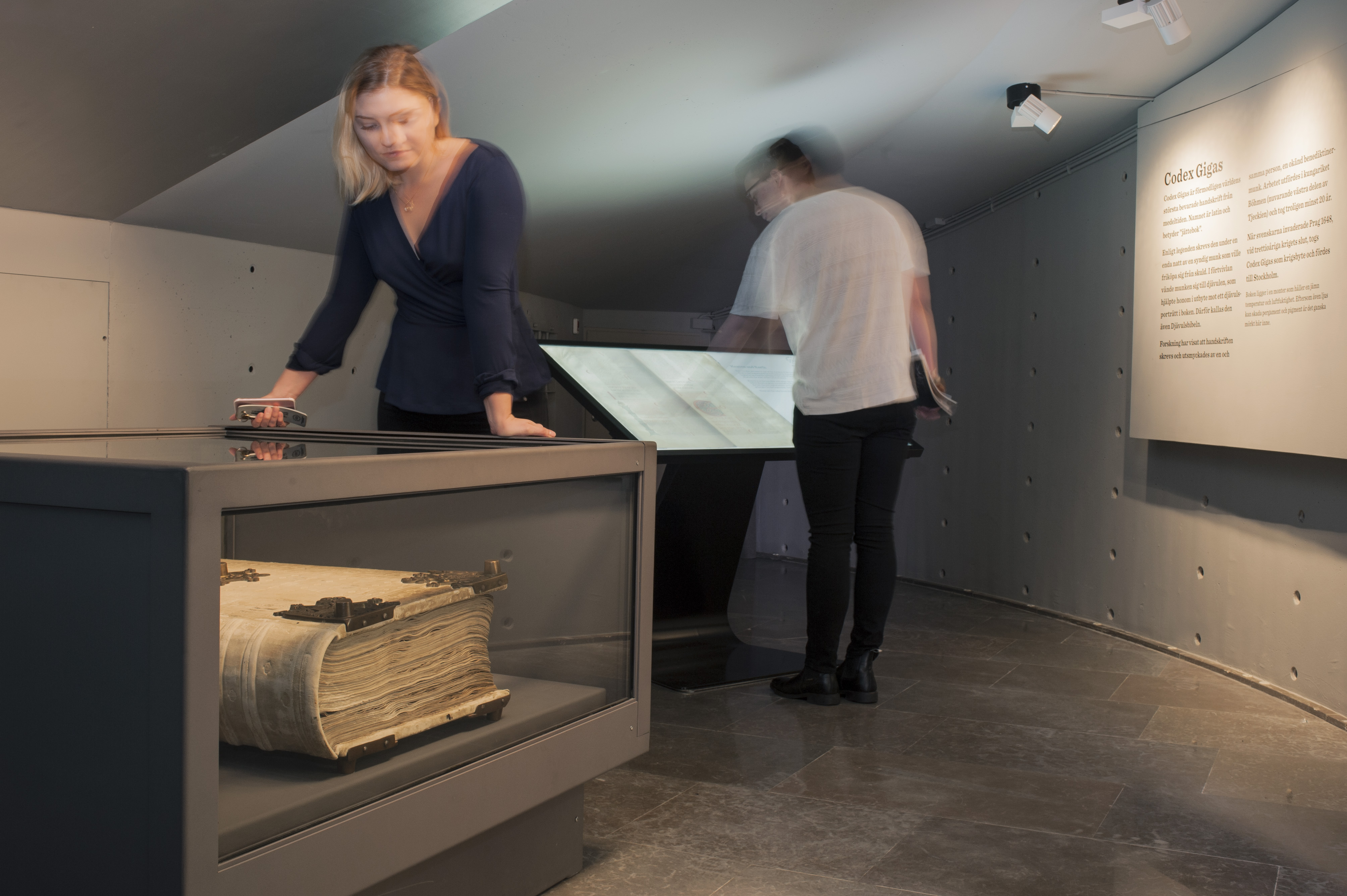 An exhibition with a big book within a glass case and two people looking around. 