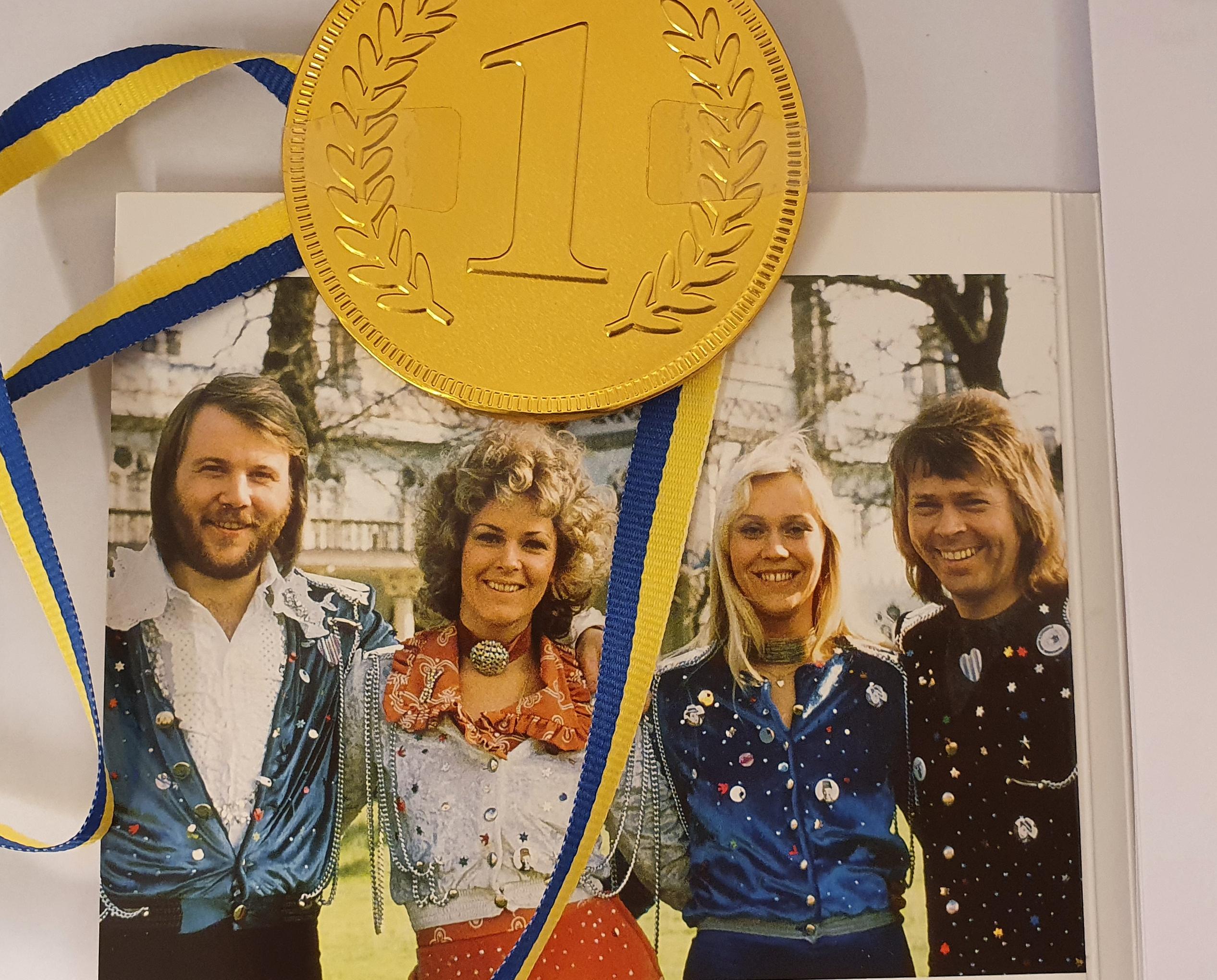 En guldmedalj med blågult band placerad över ett färgfoto med två kvinnor och två män 