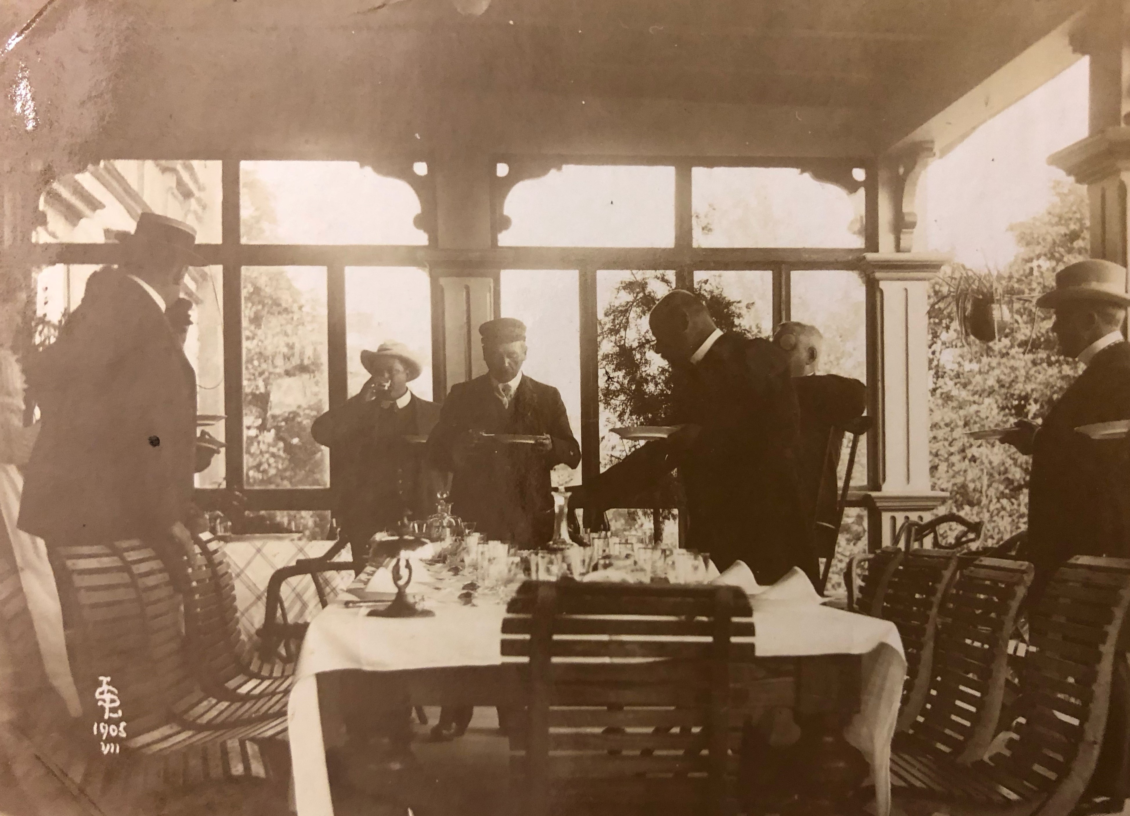 Svartvitt fotografi av ett sällskap festklädda herrar som står eller sitter runt ett stort bord på en inglasad veranda.