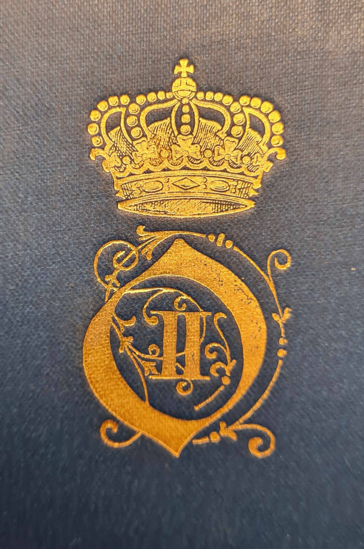 Färgfotografi av ett förgyllt monogram med kungakrona på en blå bokpärm