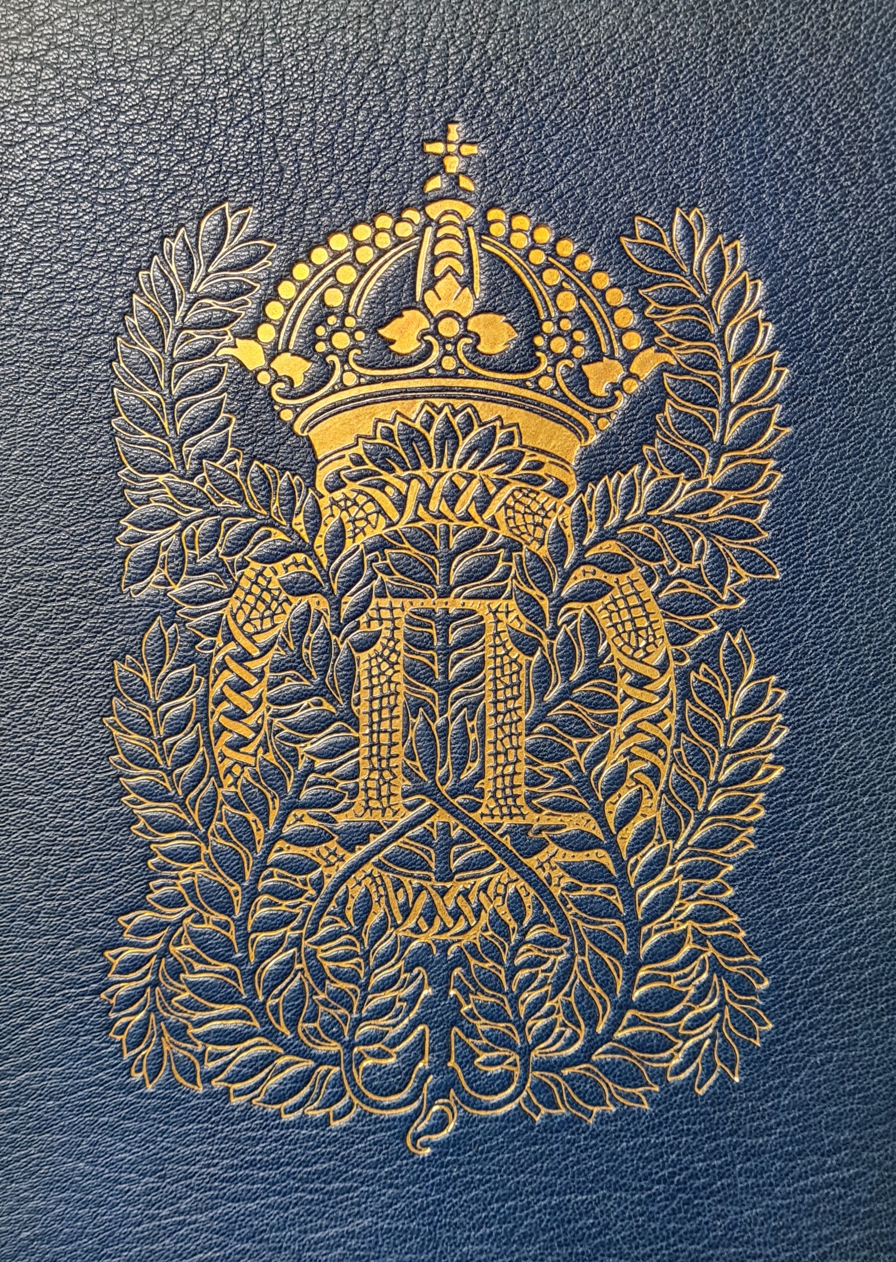 Färgfotografi av ett förgyllt monogram med kungakrona och lagerblad på en blå bokpärm