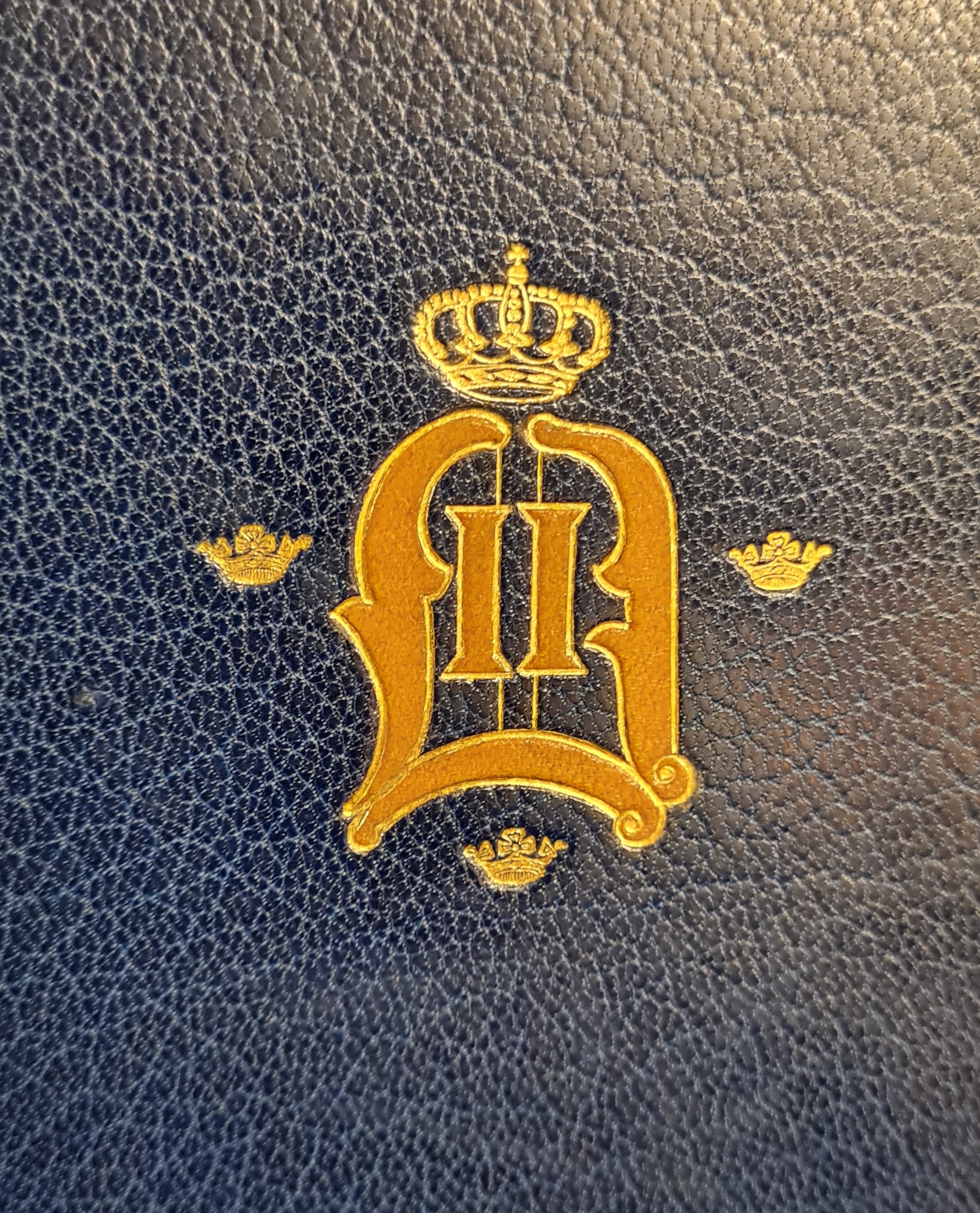 Färgfotografi av ett förgyllt monogram med kungakrona på ett blått bokband