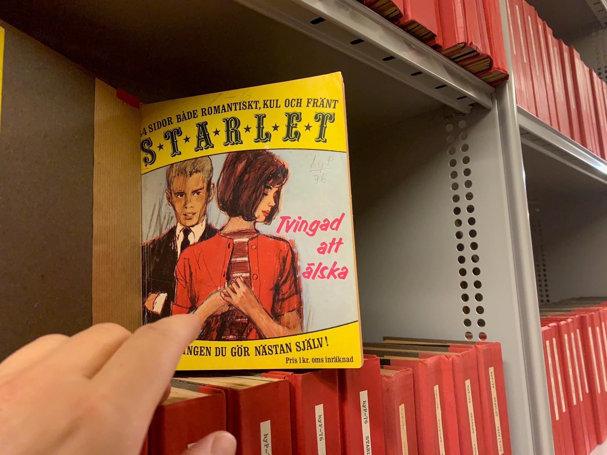 Färgfotografi av en tecknat tidskriftsomslag med ett ungt par och rubriken "Tvingad att älska", omgivet av röda bokryggar.