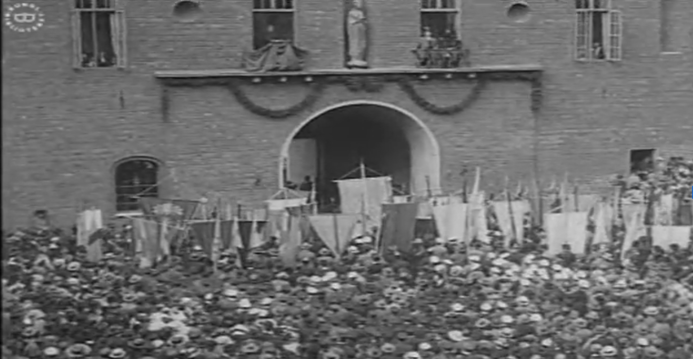 Svartvit bild på folkmassa framför en stor byggnad. Flera bär hatt och håller upp flaggor.