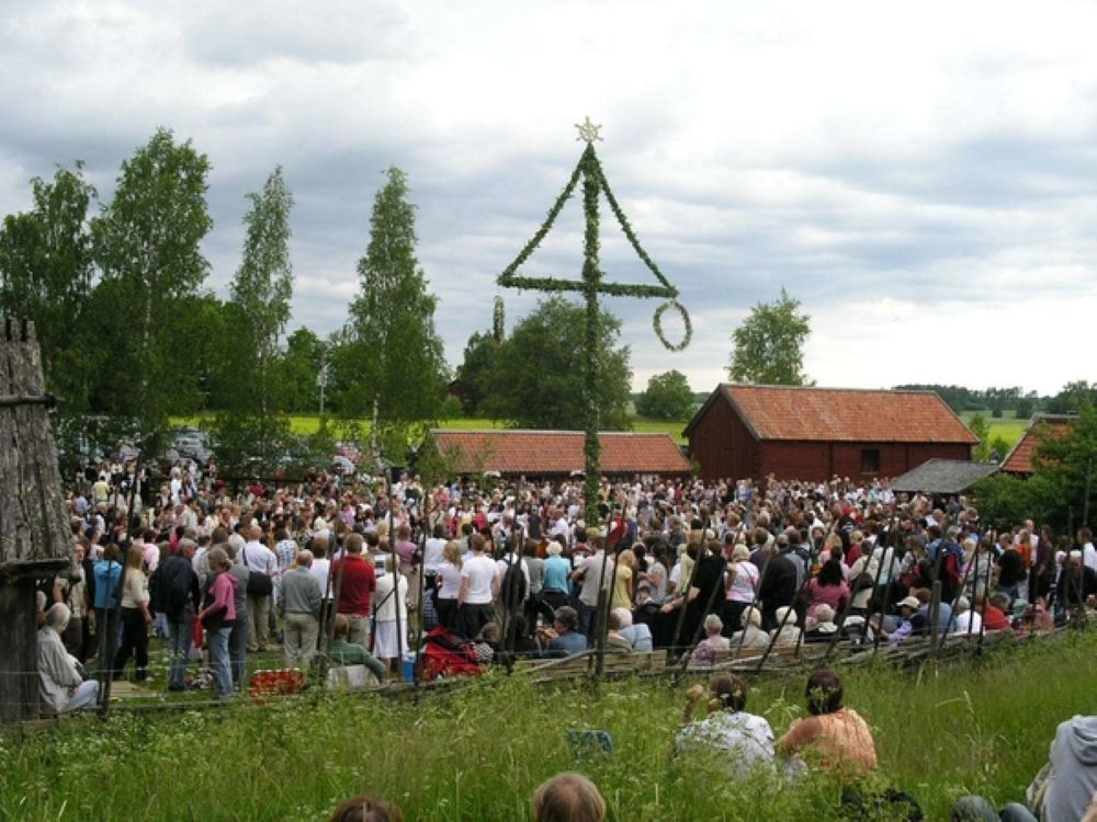 Stor samling av människor som dansar kring en stång. Röda hus i bakgrunden.