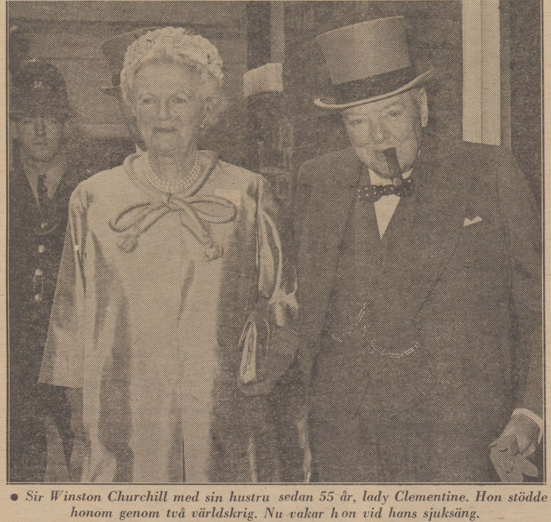 Gulnat tidningsfotografi, dam i sidenklänning och herre i kostym, hatt och med cigarr. Text: Sir Winston Churchill med sin hustru.