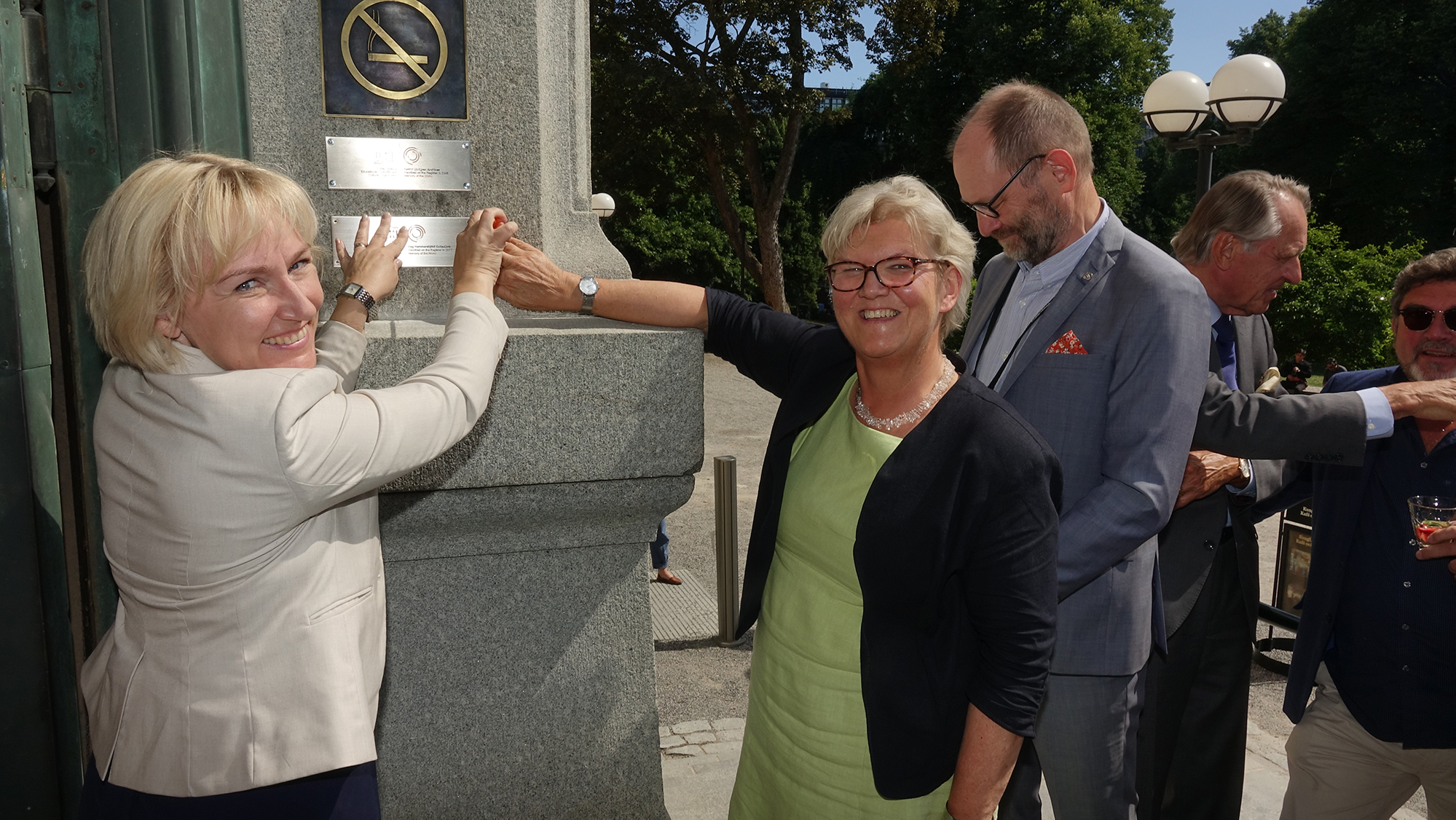 Två kvinnor ler och håller sina händer på en skylt som spikats upp på en pelare. I bakgrunden syns träd i en park.