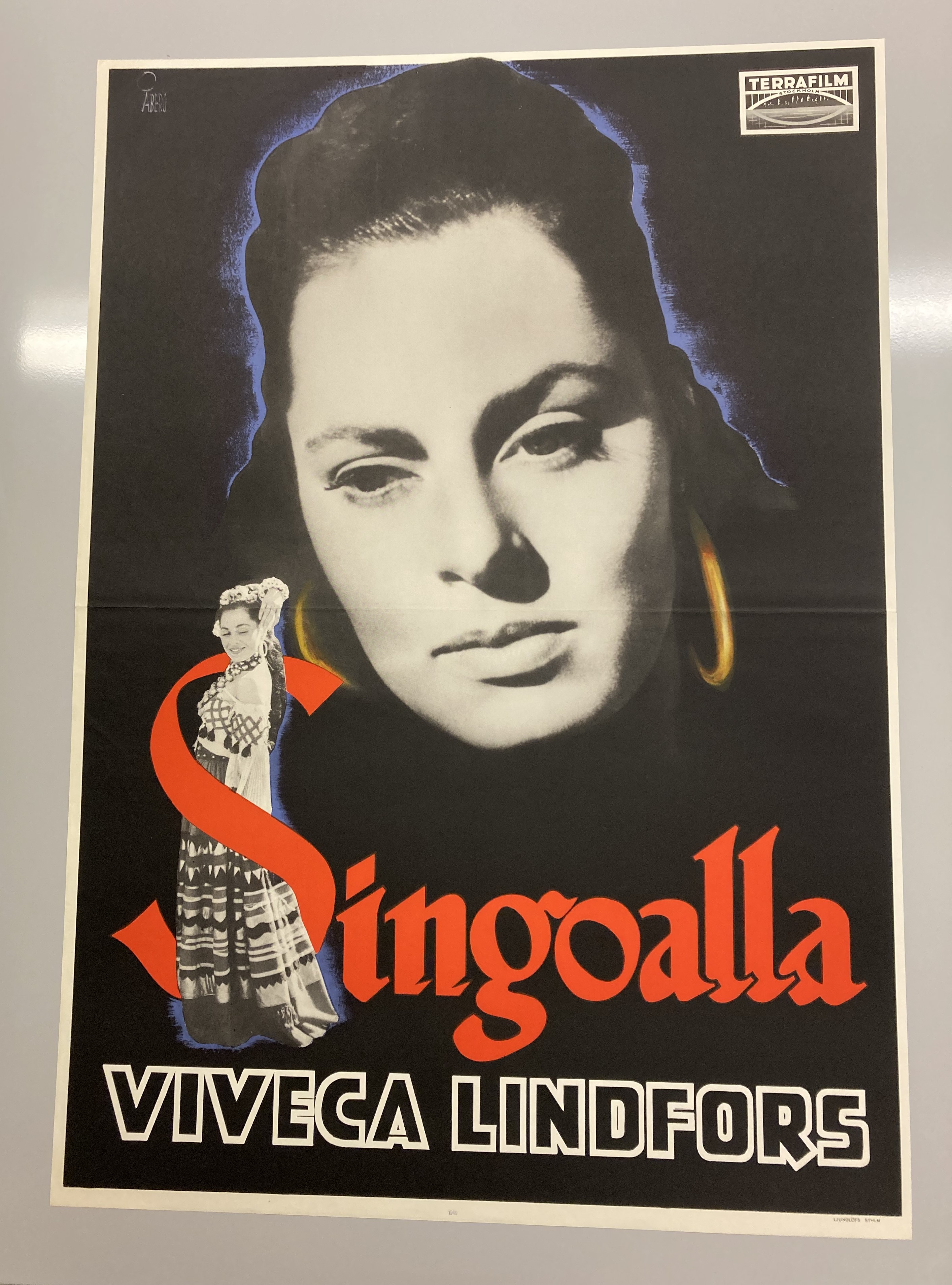Färgfoto av en filmafffisch med en stor närbild på en vacker ung kvinna med stora ringar i öronen. Nertill en mindre svartvit bild av samma kvinna i helfigur iklädd romska kläder, stående inuti ett rött S som inleder titeln Singoalla.