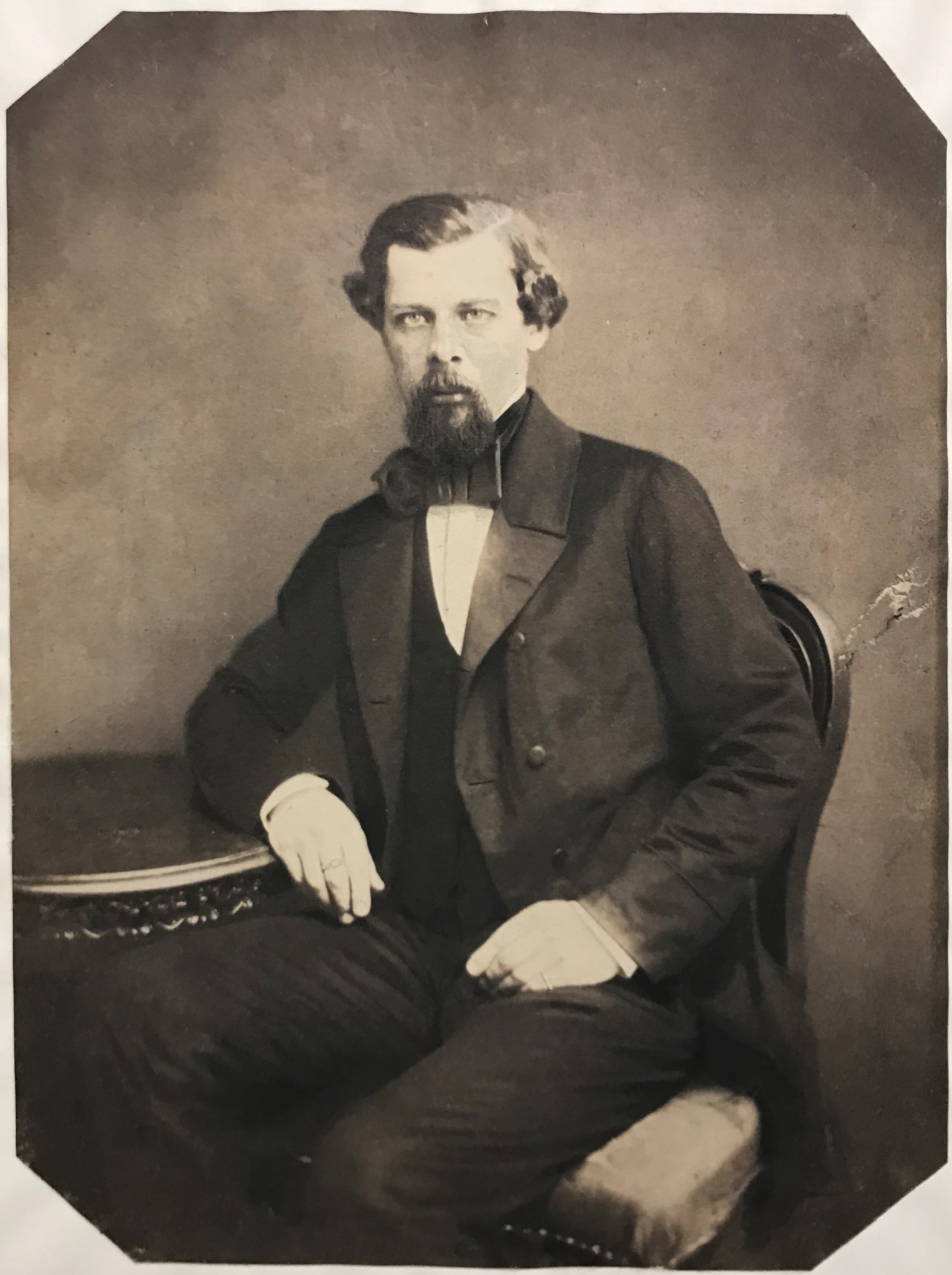 Svartvitt fotografi av en kostymklädd ung man med mustasch och kort skägg sittande vid ett bord.
