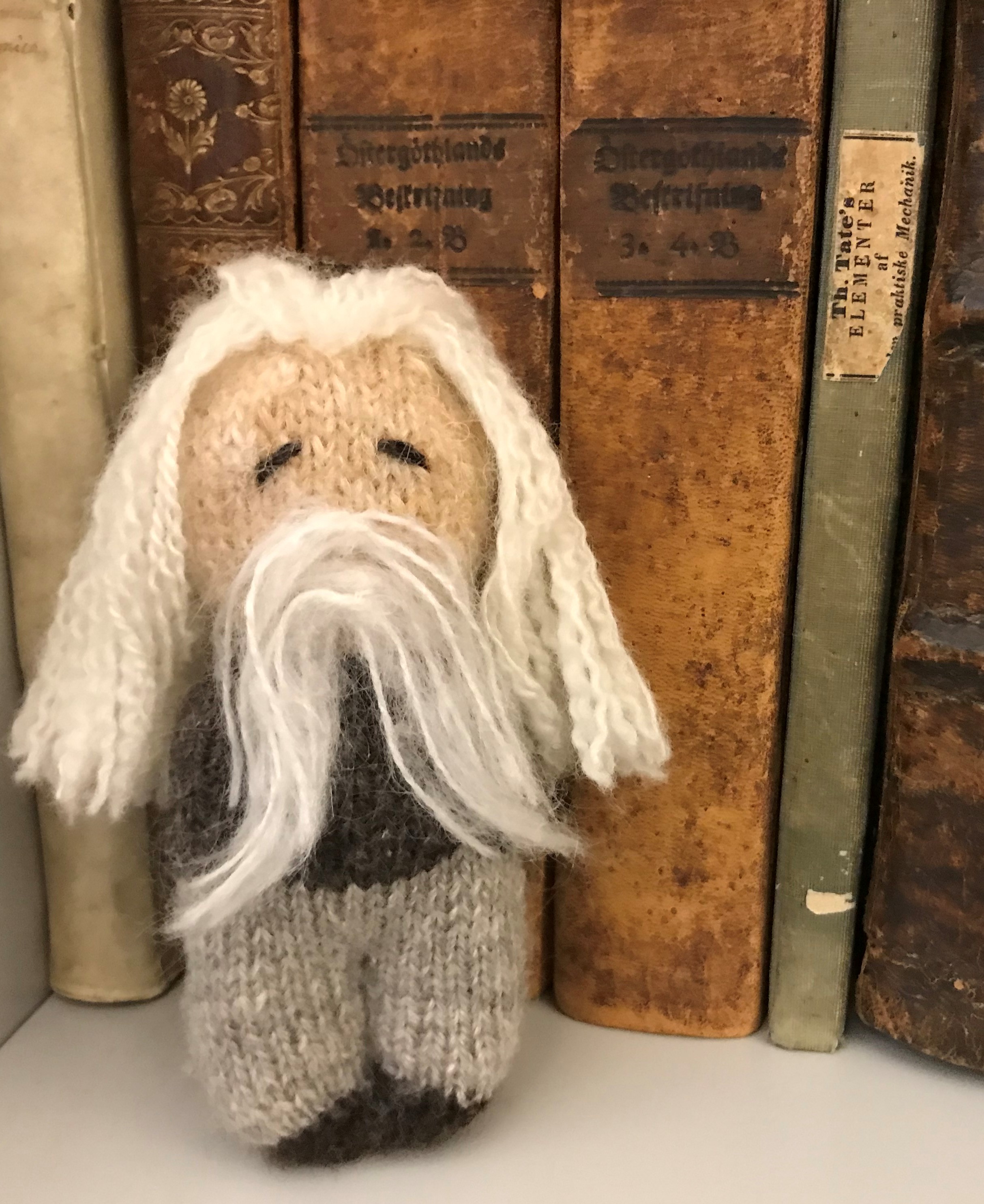 En stickad liten docka med långt hår och skägg står på en hylla lutad mot ett par gamla bokryggar.