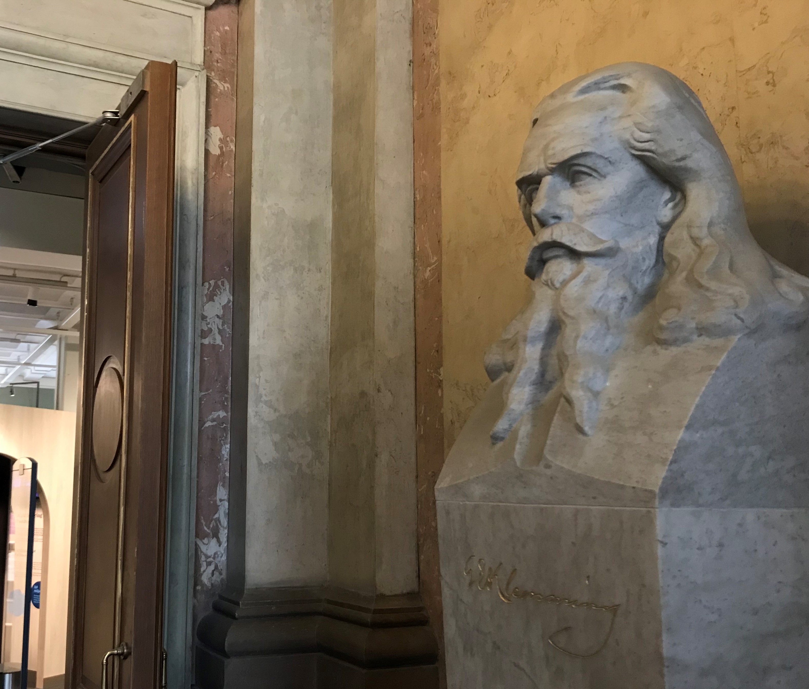 Färgfotografi av en marmorbyst föreställande en man med långt hår och långt skägg, placerad framför en öppen dörr.