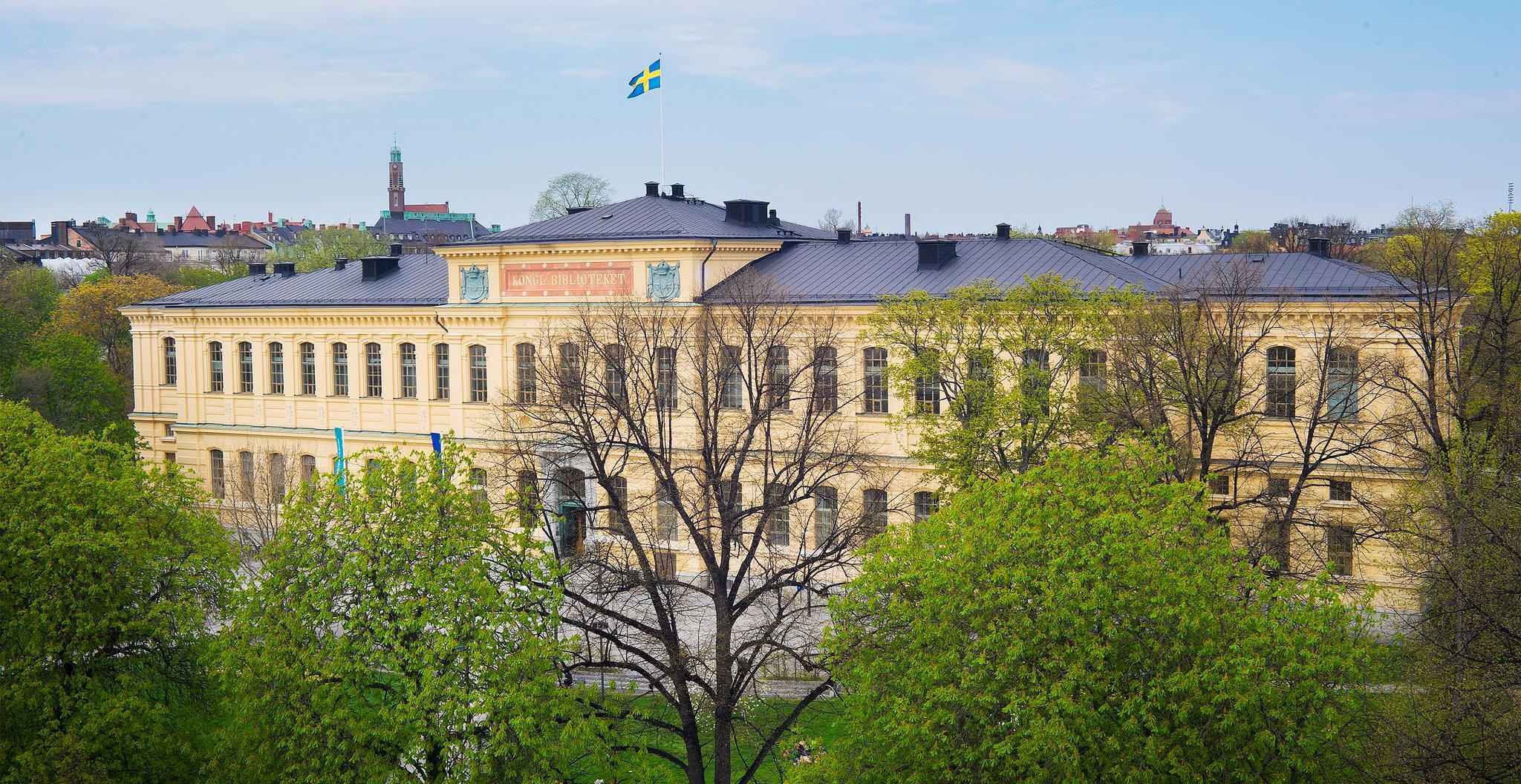 Kungliga bibliotekets gula byggnad sedd uppifrån. Från tacket vajar en svensk flagga och runt omkring finns parken Humlegården.