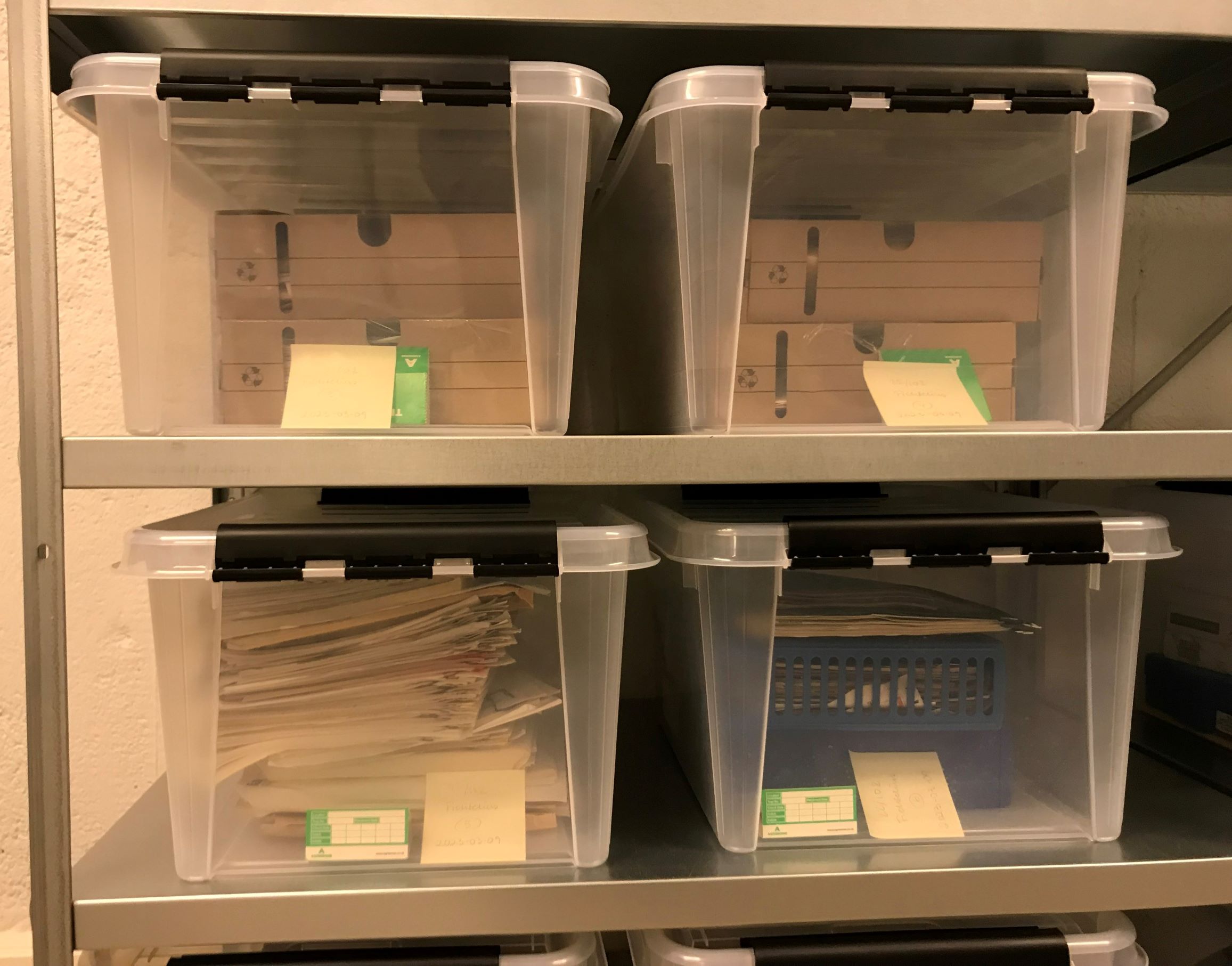 Två hyllplan med fyra genomskinliga plastbackar som innehåller kartonger och dokument. I varje plastback ligger en grön trekantig insektsfälla i papp.