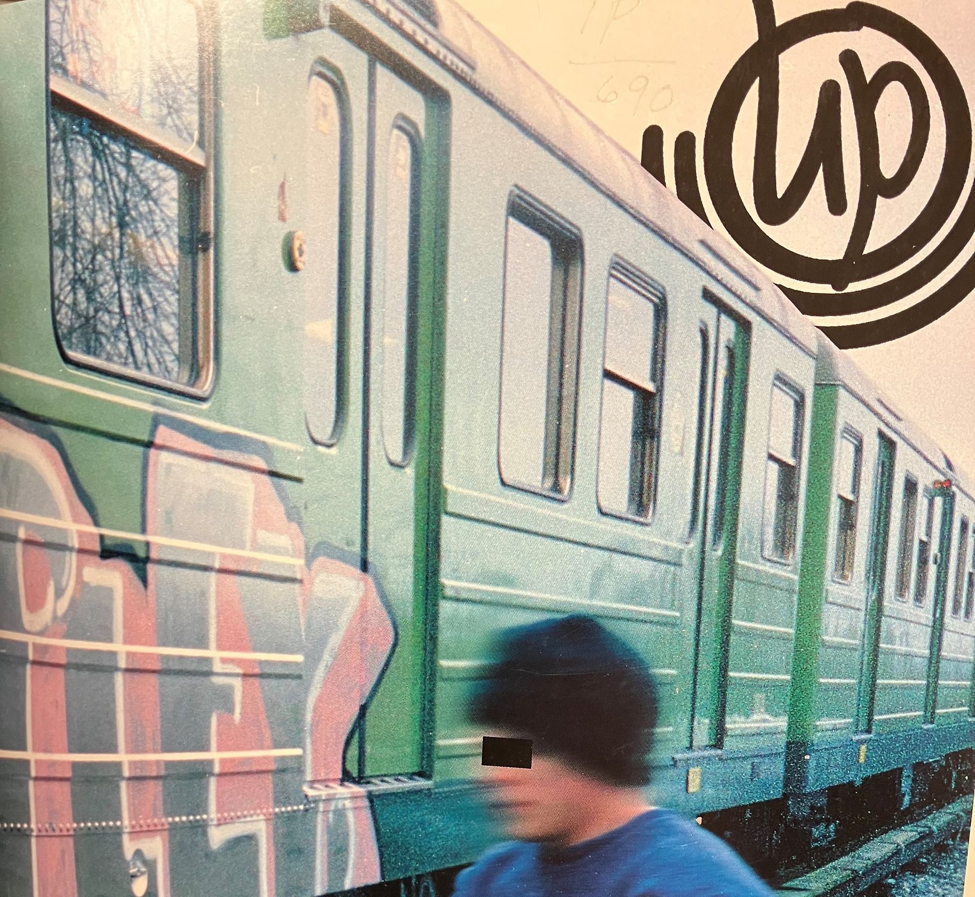 Färgbild föreställande delar av en grön tunnelbanevagn med graffiti. Bredvid syns huvudet på en ung person med svart sotarmössa, ögonen maskerade av fyrkant. Logotypen för UP står skrivet uppe i högra hörnet.