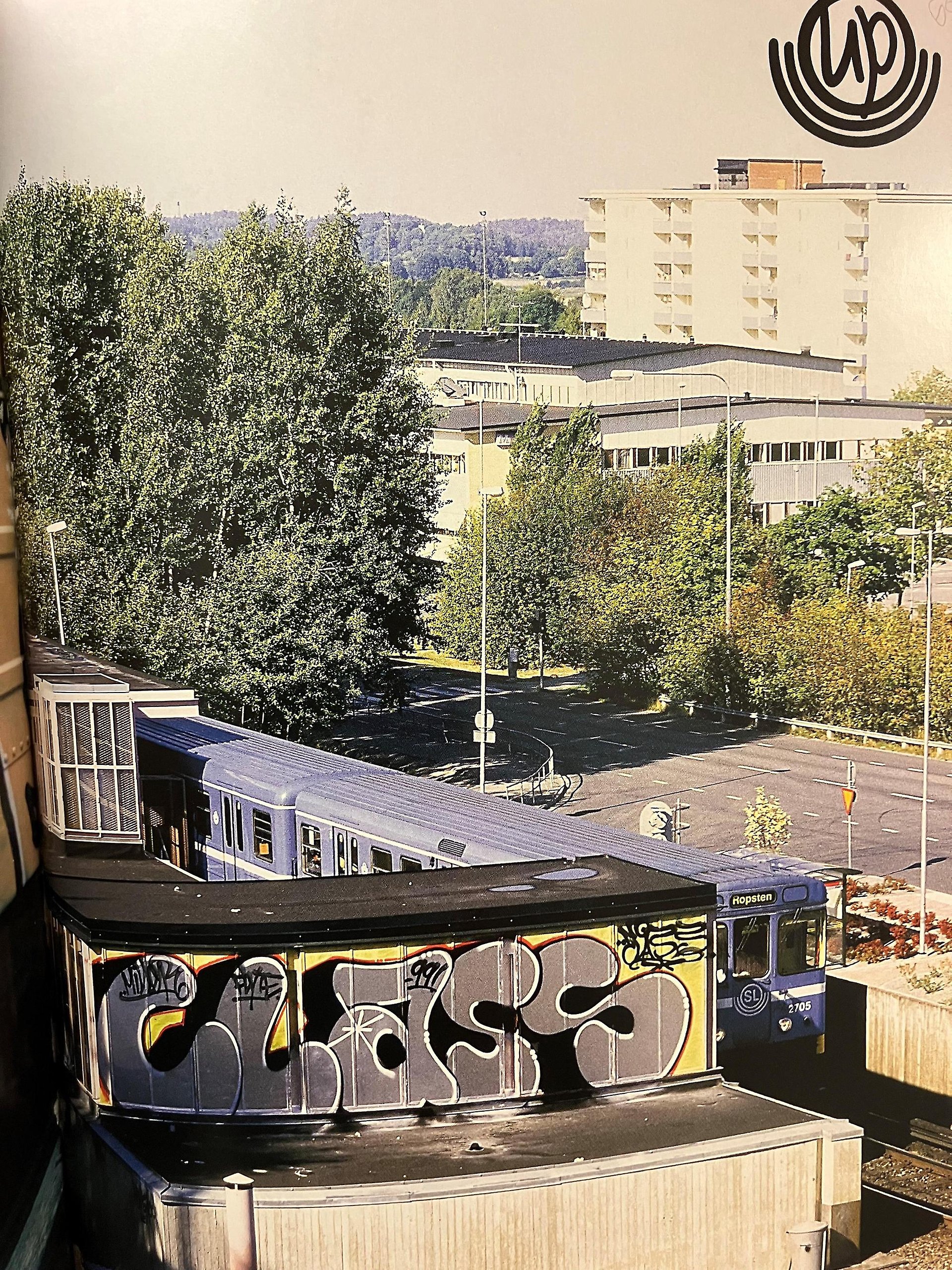 Vy i färg med tunnelbanetåg, höghus och träd. I förgrunden en vägg med graffiti och texten class. Logotypen UP syns i övre högra hörnet.