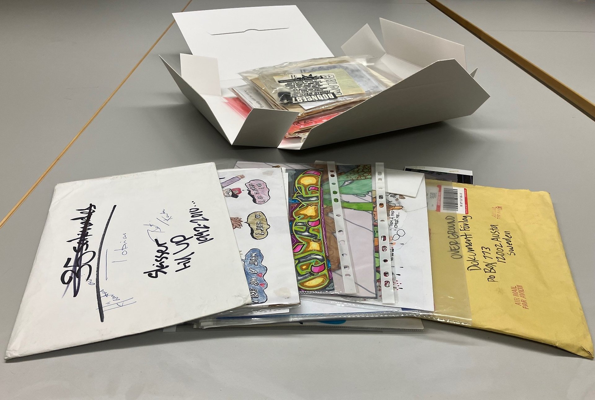 Färgfoto av en öppnad arkivkapsel full med material i plastfickor, framför den ligger kuvert och plastfickor med färgglada graffitiskisser.