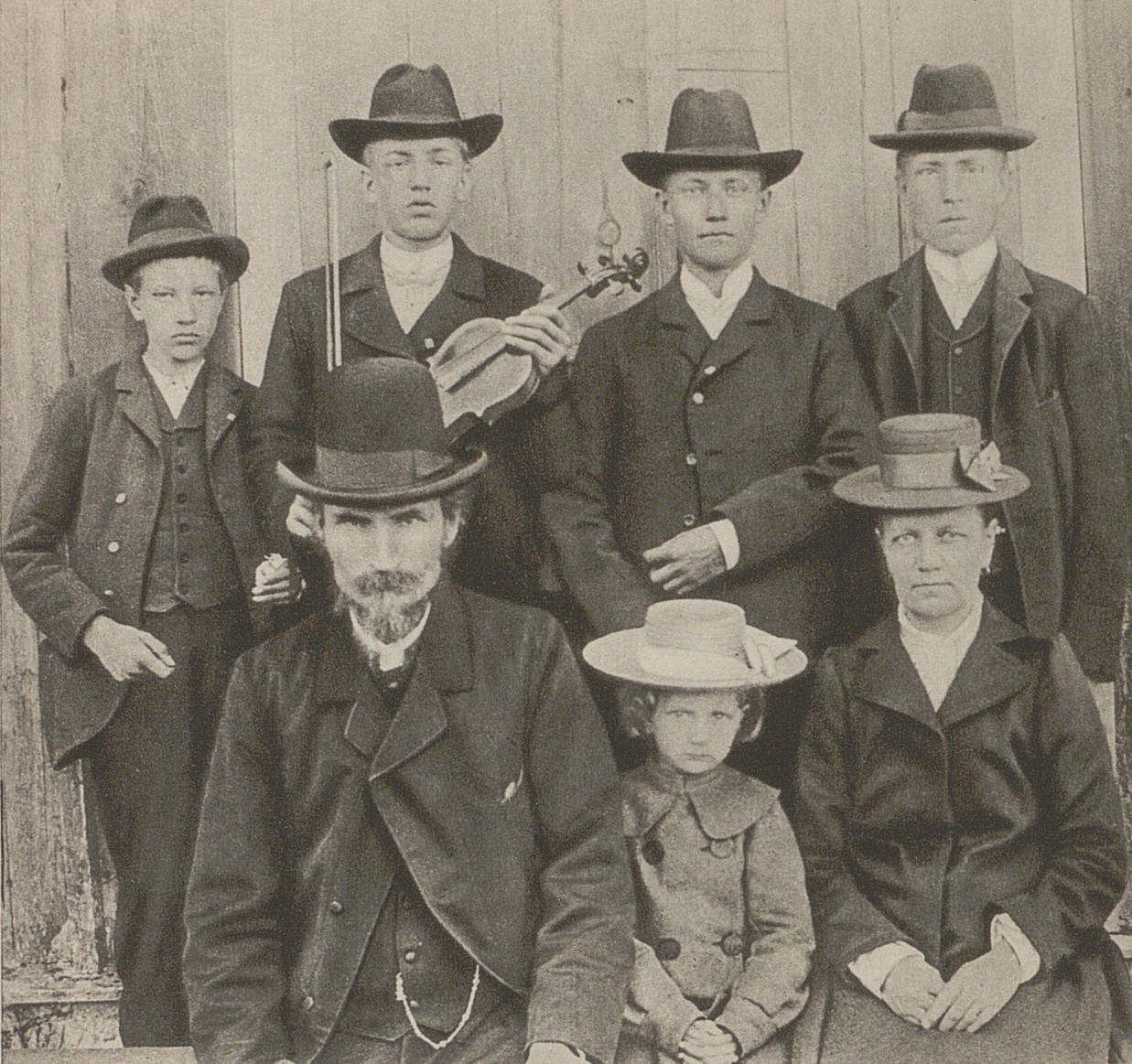 Svartvitt foto på familj, fyra pojkar längst bak i varierande åldrar. Längst fram föräldrar och liten dotter, alla har hatt och ser allvarliga ut.