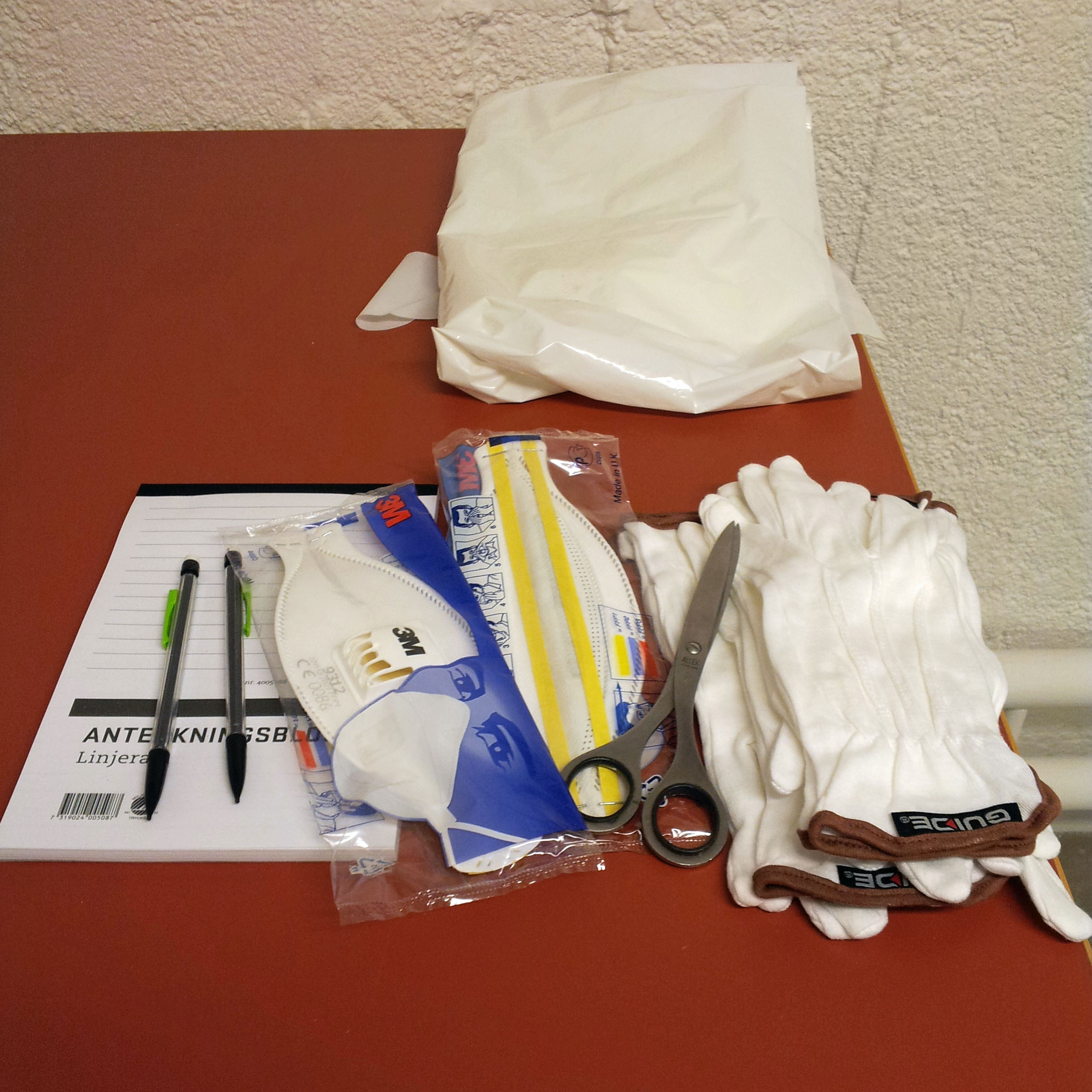Vantar, andningsskydd, plastförkläde, anteckningsblock och pennor ligger på ett bord.