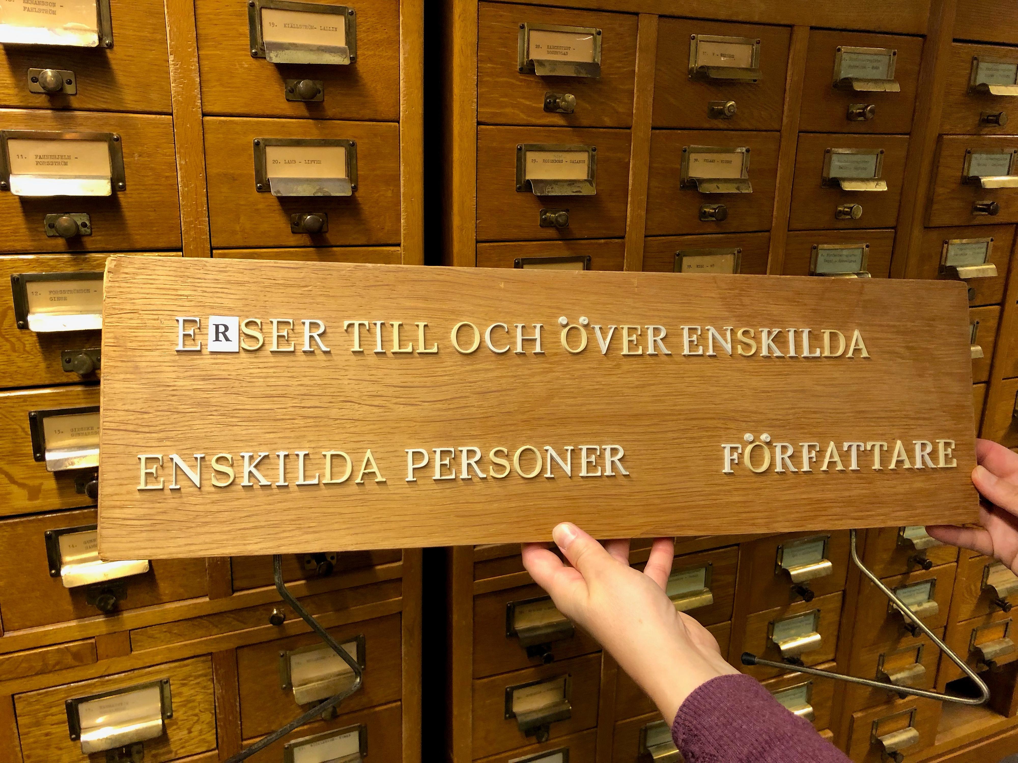 Två händer håller upp en äldre skylt i trä framför en kortkatalog. Texten på skylten lyder Verser till och över enskilda, enskilda personer författare. Någon bokstav saknas.