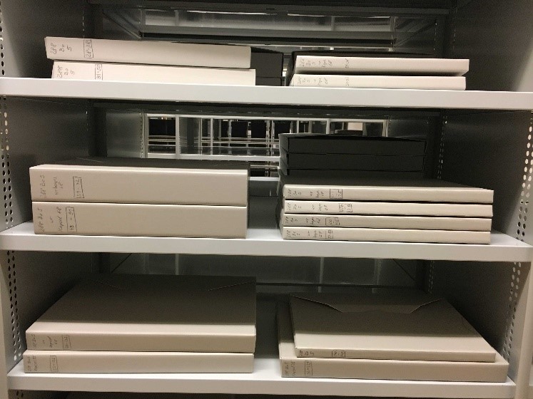 Färgfoto av tre hyllplan där det ligger travar med grå arkivkapslar.