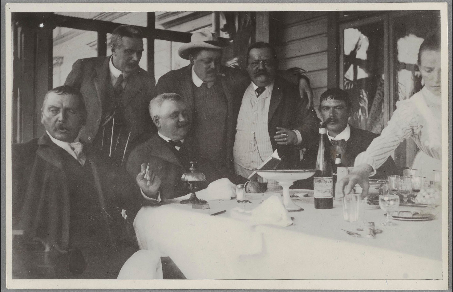 Ett svartvitt fotografi med sex män och en kvinna vid ett halvt avdukat bord. Männen sjunger.