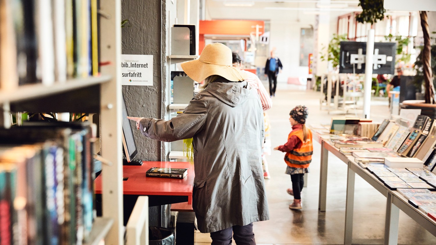 En äldre person i hatt och ett barn med orange väst i en biblioteksmiljö.