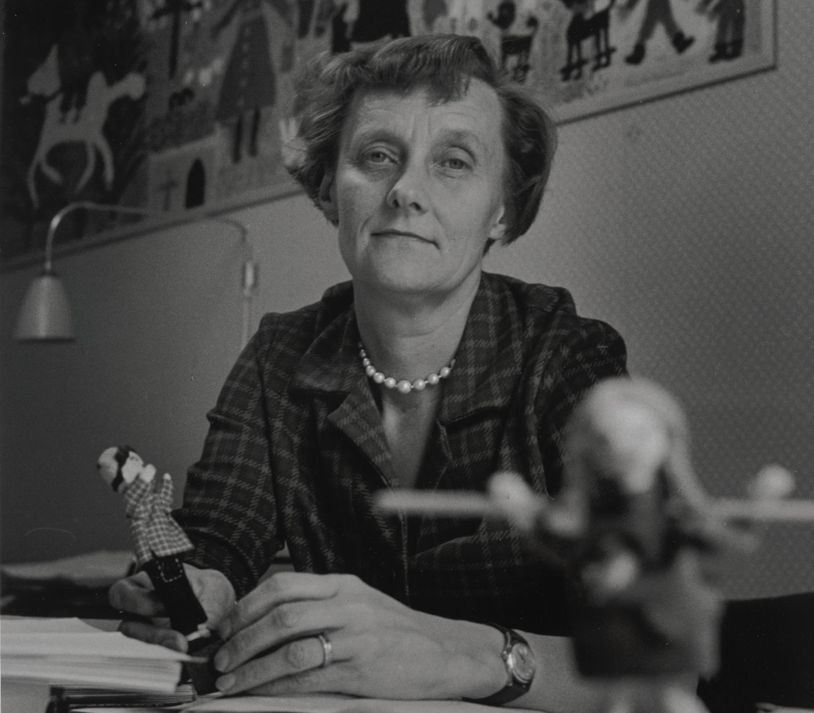 Svartvitt fotografi av Astrid Lindgren som sitter vid ett skrivbord. I ena handen håller hon en docka som ser ut som Emil i Lönneberga, i bildens förgrund skymtar en docka som ser ut som Pippi Långstrump.