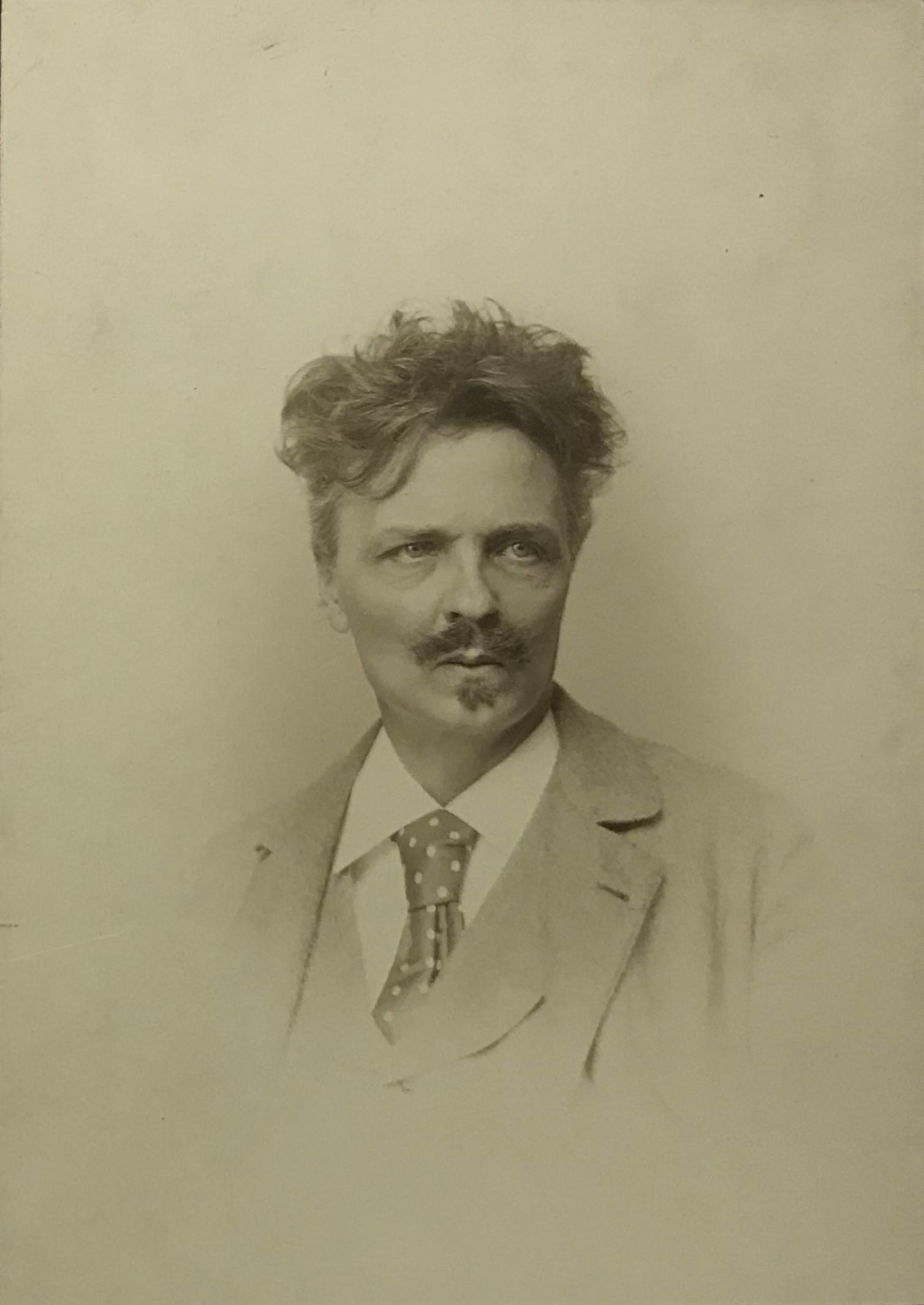 Svartvitt fotografi i halvfigur av en man med mustasch och yvigt hår.