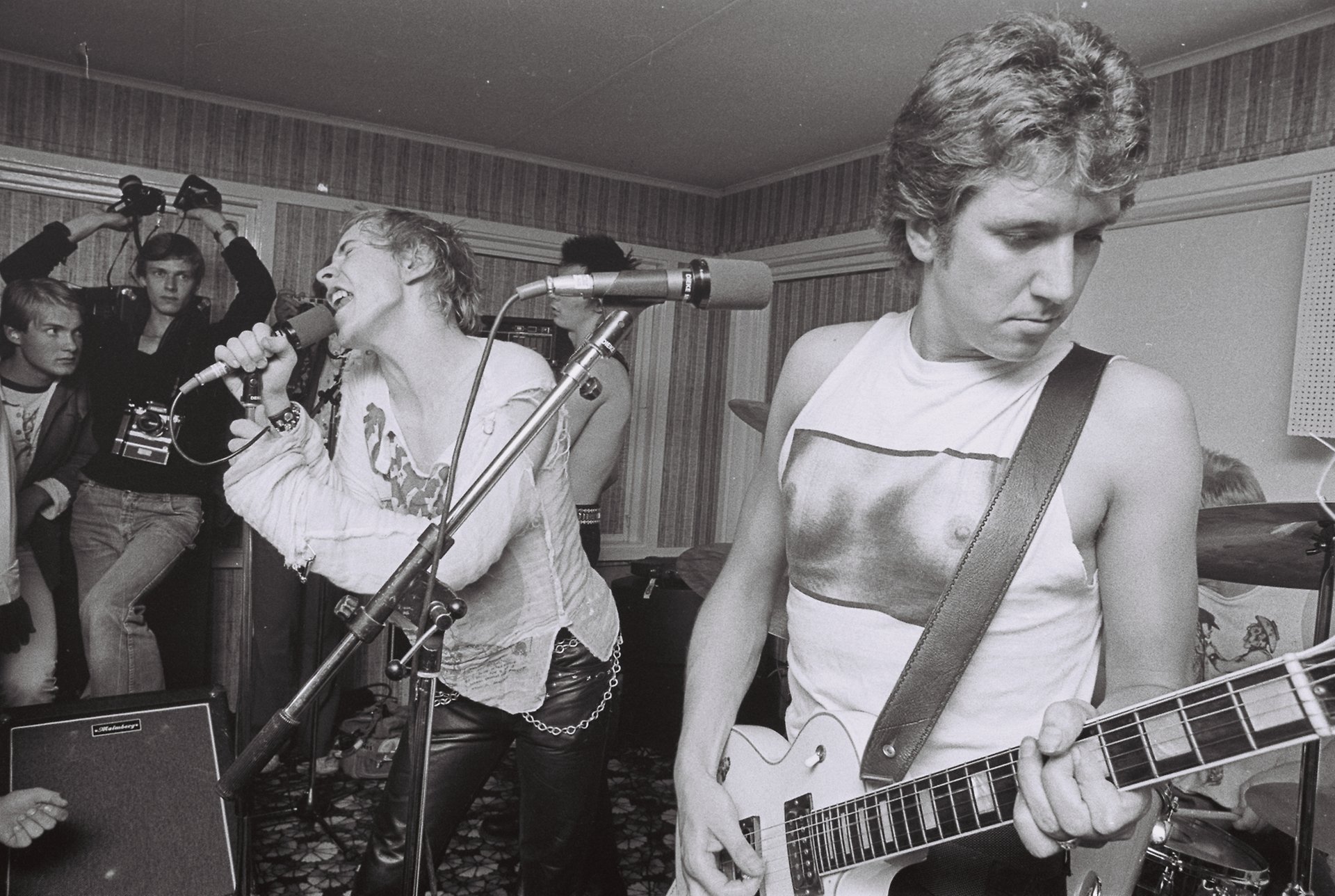 Svartvitt fotografi av en ung man klädd i läderbyxor, nitar och en söndertrasad tröja som sjunger i mikrofon bredvid en man som spelar gitarr iförd en ärmlös t-shirt med tryckta kvinnobröst.