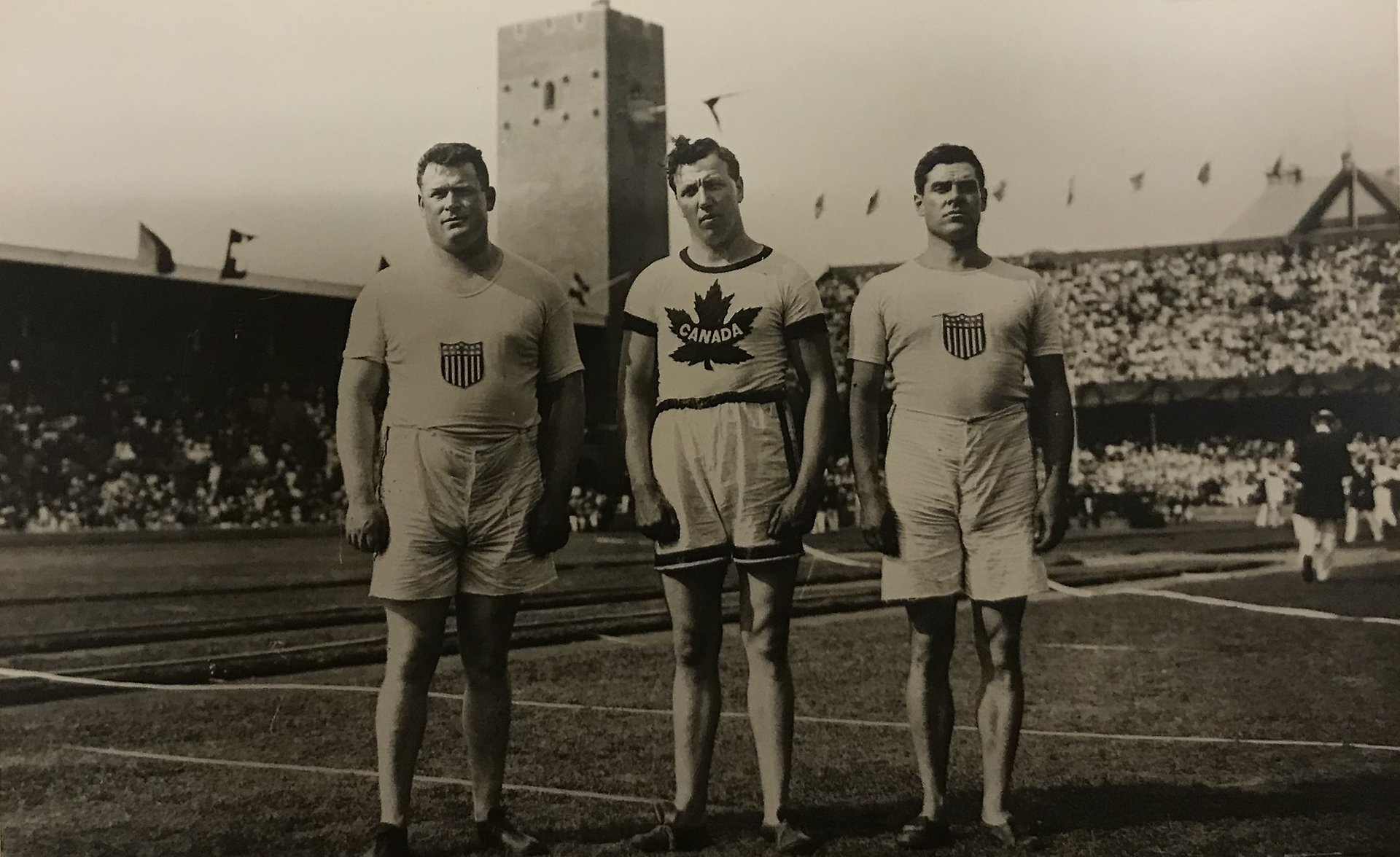 Svartvitt fotografi tre unga män klädda i shorts och t-shirts med amerikanska och kanadensiska emblem stående på gräsmattan på Stockholms stadion.