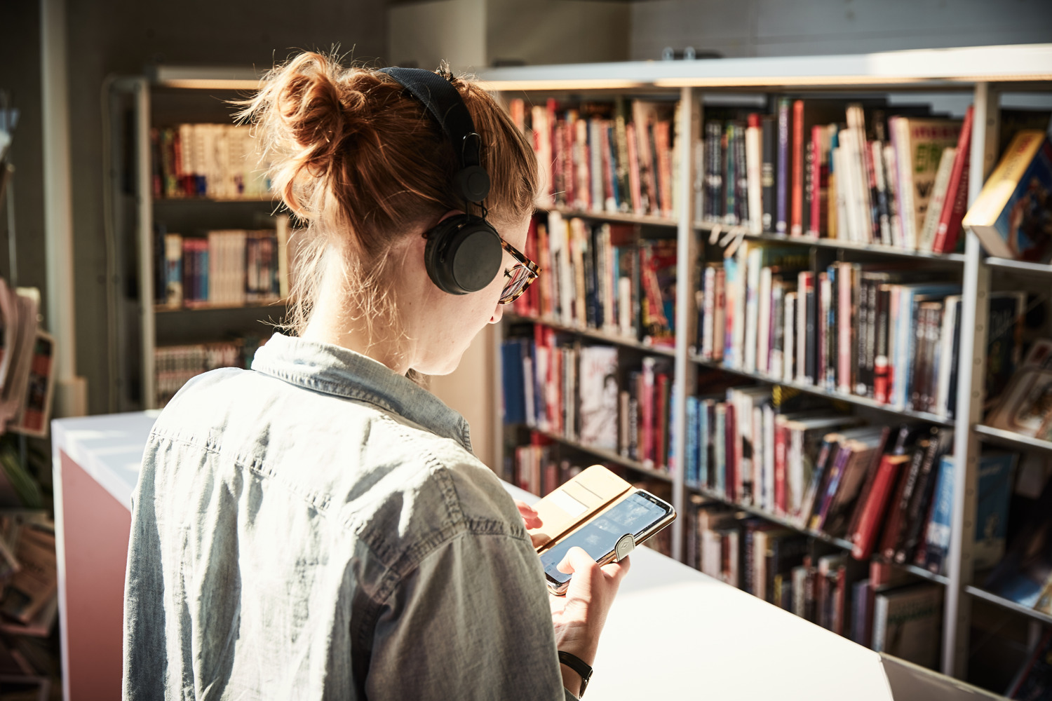 Foto av en person snett bakifrån, personen har på sig hörlurar, håller i och tittar på en smartphone i biblioteksmiljö