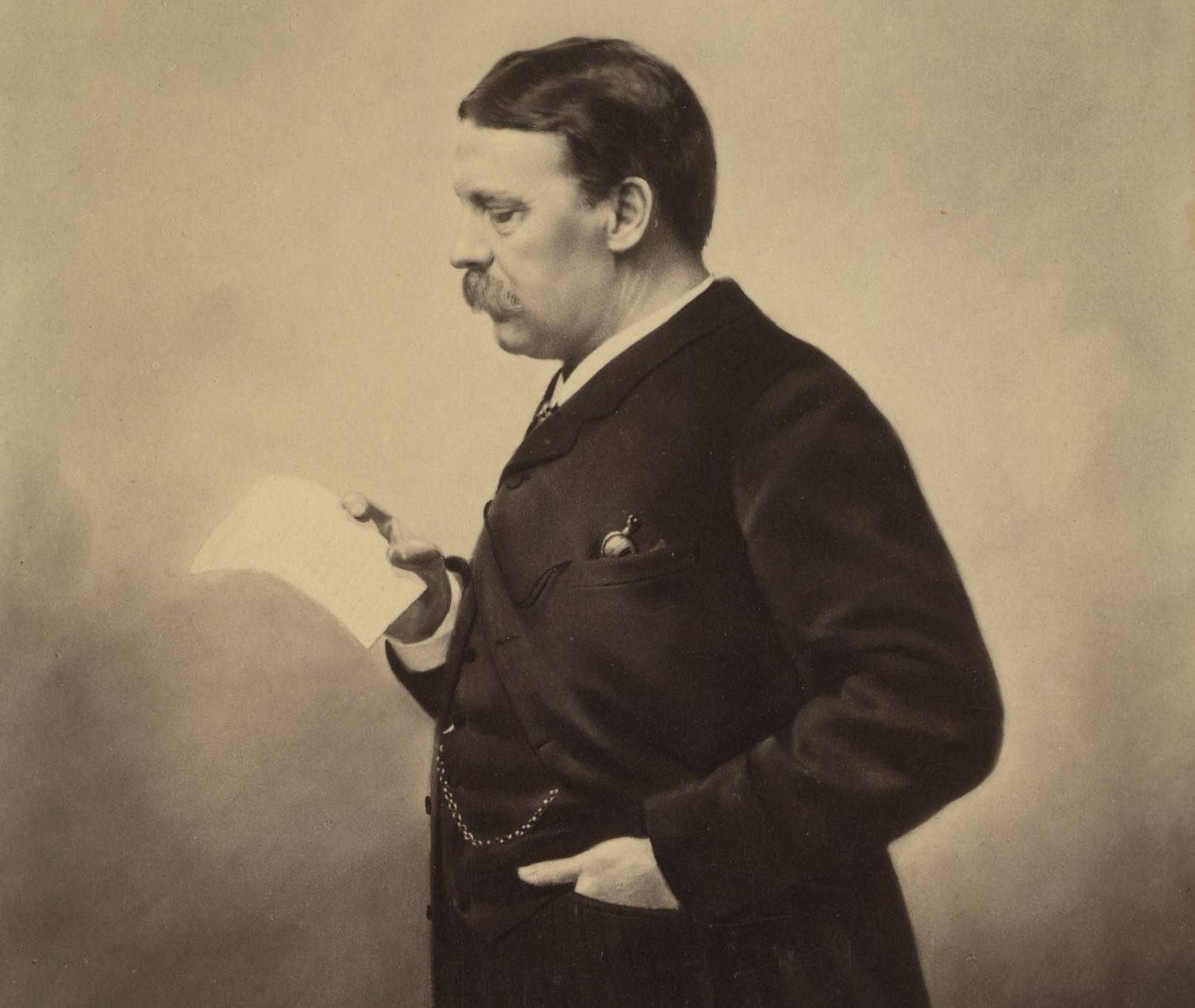Svartvitt fotografi av en medelåldersman med mustasch avbildad i profil som läser ett papper han håller i höger hand, den vänstra handen har han i en byxficka.