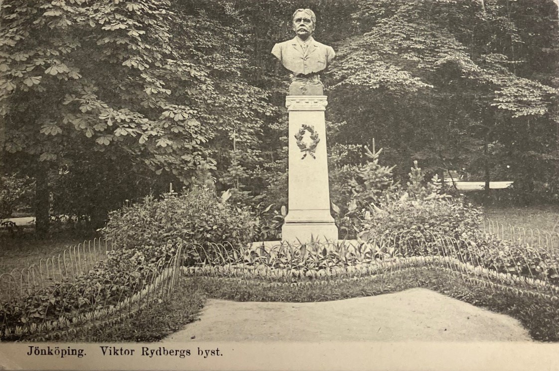 Svartvitt vykort föreställande en parkmiljö med en byst av en man med mustasch på en pelare prydd med en lagerkrans.