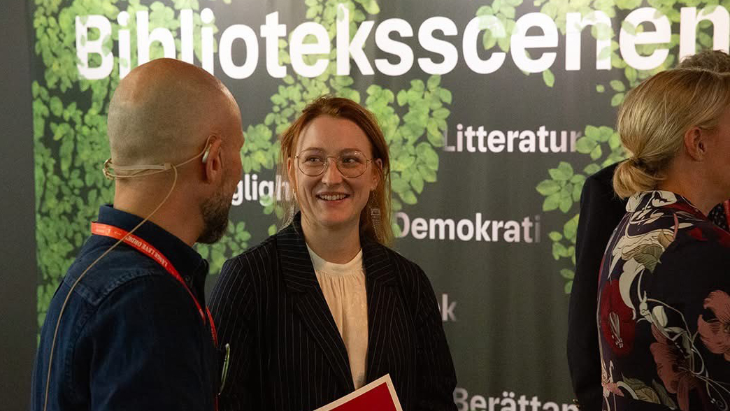 En man och en kvinna står och samtalar framför en grön bakgrund med texten Biblioteksscenen