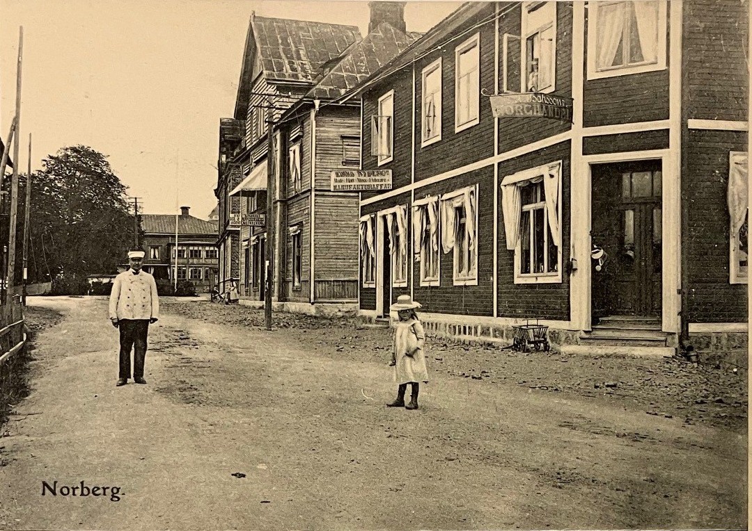 Svartvitt vykort med stadsbild. En man i kostym och stråhatt och en flicka i klänning står på en grusväg kantad av trähus.
