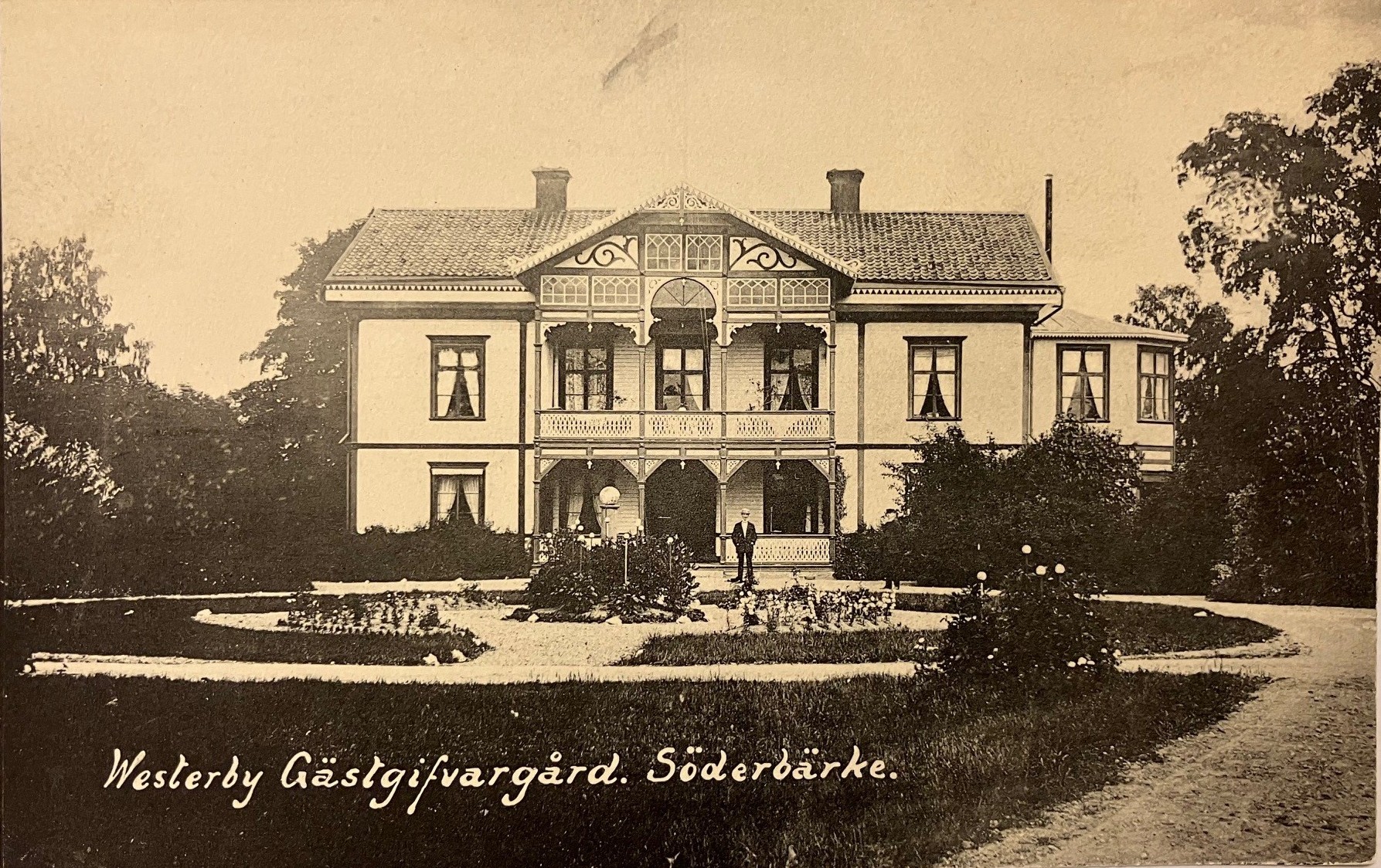 Svartvitt vykort på trädgård och trähus med konstnärligt utförda snickerier i mittpartiet.