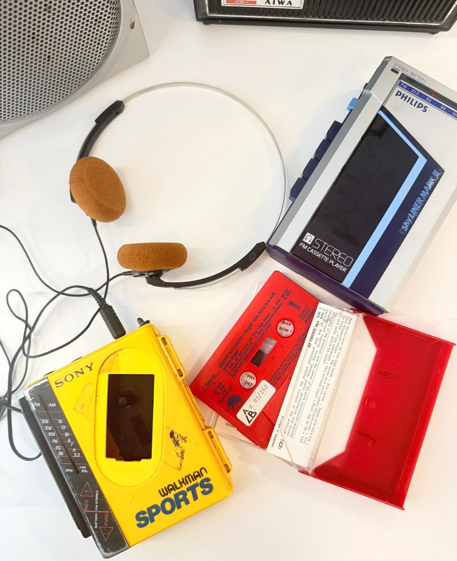En gul Sony Walkman ligger på ett bord tillsammans med en bärbar kassettspelare från Philips, ett par hörlurar och ett kassettband.