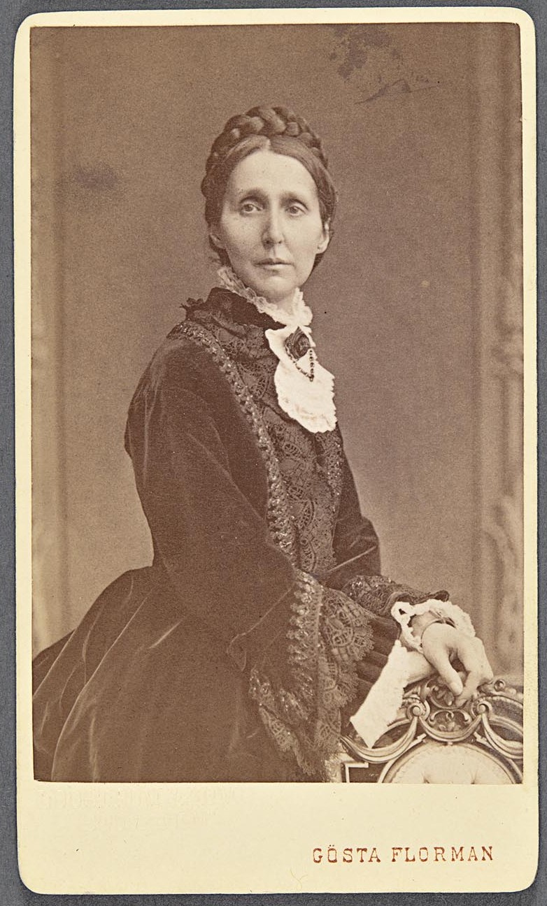 Svartvitt porträttfoto av kvinna klädd i sammetsdräkt och broderad blus. Hon har håret i en fläta på huvudet.