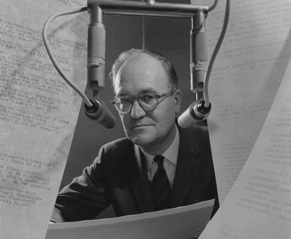 Fotografi av en tunnhårig man med glasögon bakom två mikrofoner och maskinskrivna papper.