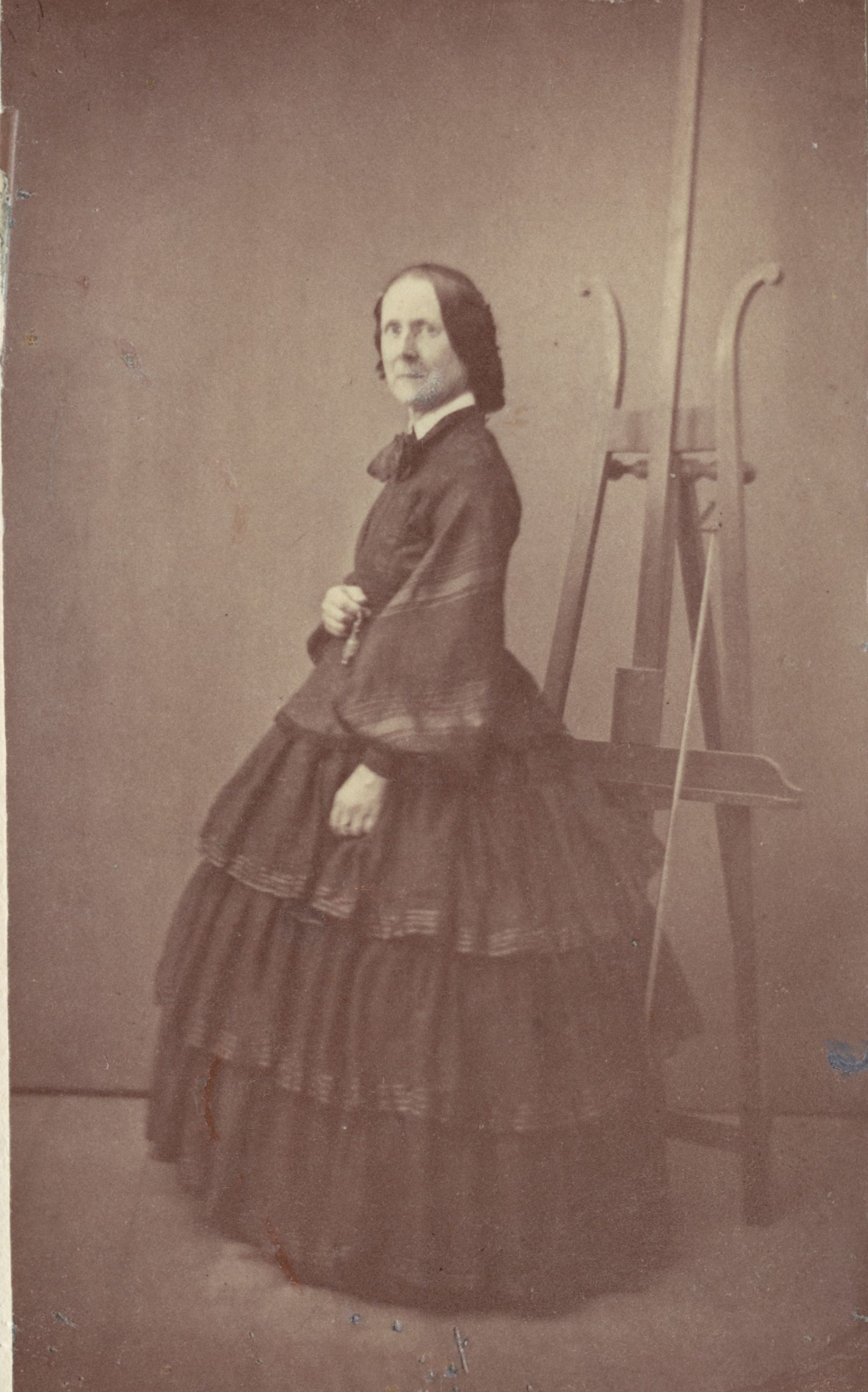 Svartvitt fotografi av en äldre kvinna i mörk klänning stående framför ett staffli.