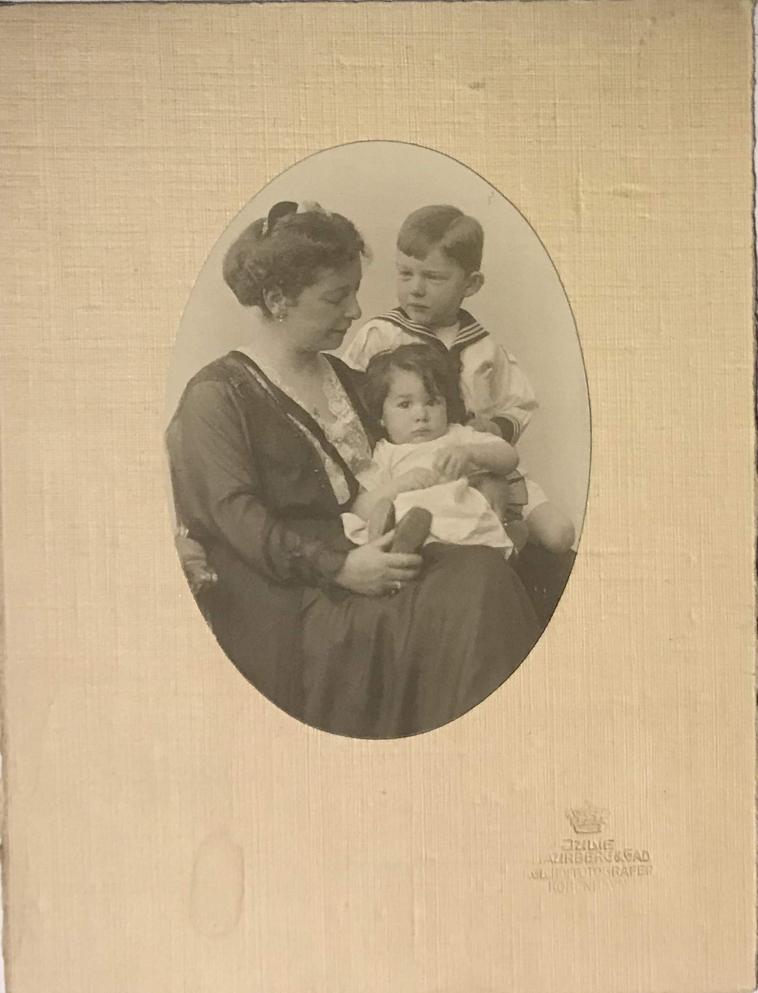 Svartvitt fotografi med en sittande kvinna som håller ett litet barn i famnen. Bredvid sitter en pojke i sjömanskostym.