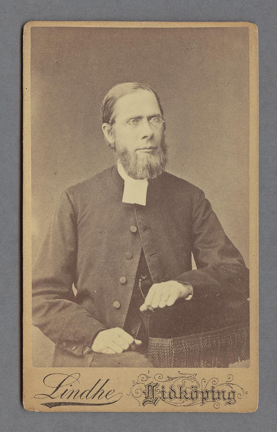 Porträtt av Johannes Granqvist iklädd rock och prästkrage. Hans sitter ner och hans högra arm är placerat i knät medans den vänstra vilar på ett tygbeklätt armstöd med fransar.