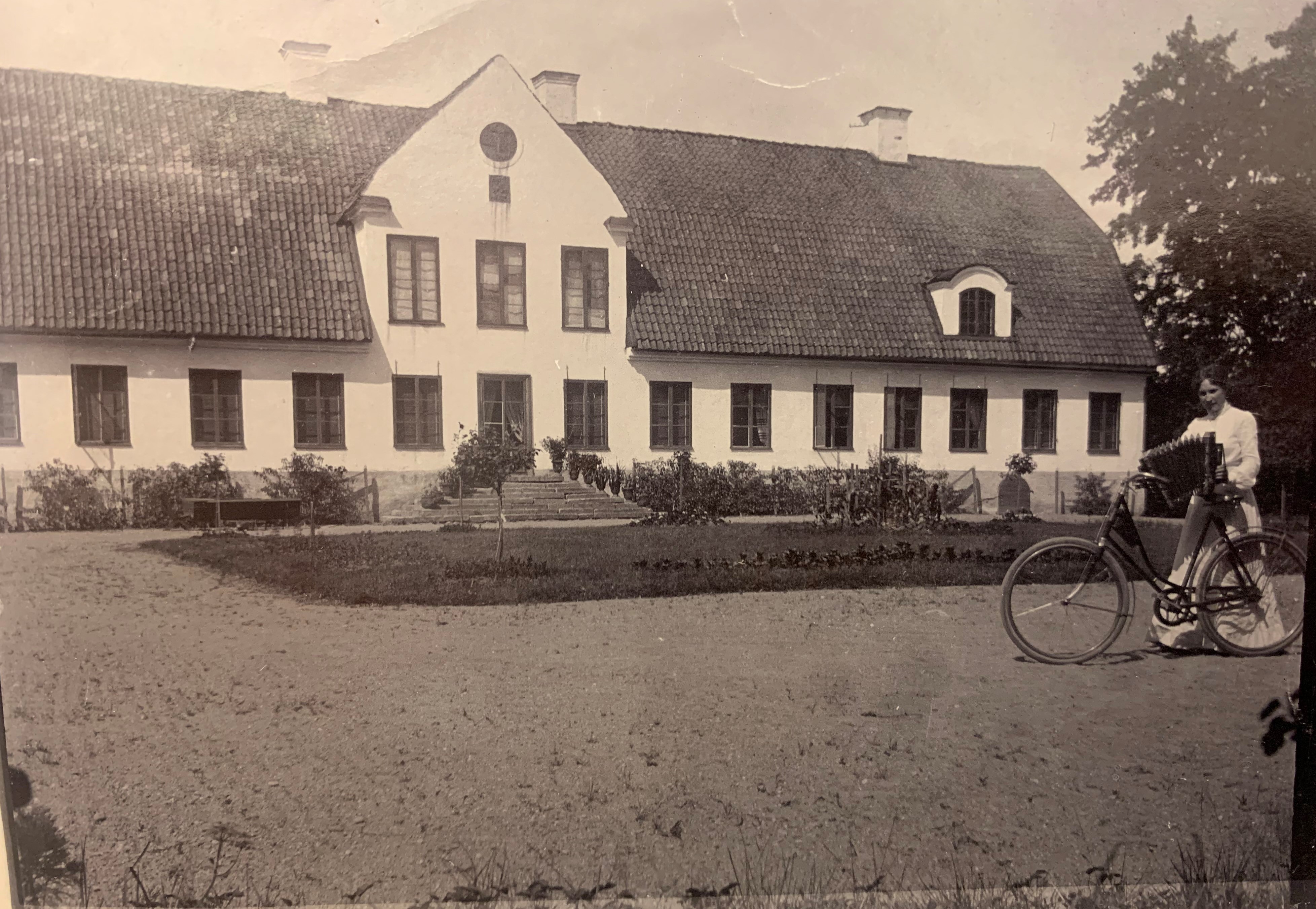 Svartvitt foto av gården Säby. I förgrunden en kvinna med cykel