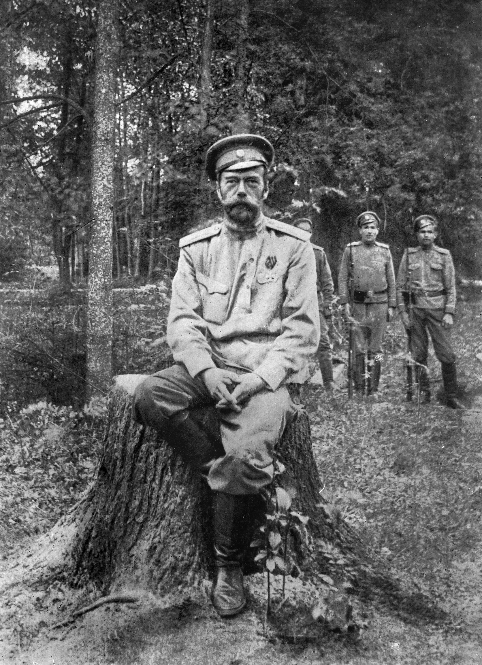 Svartvitt fotografi av en man i uniform med tillhörande hatt. Han sitter på en stubbe i skogen och tittar med allvarlig min mot kameran. I bakgrunden syns tre andra män i uniform.