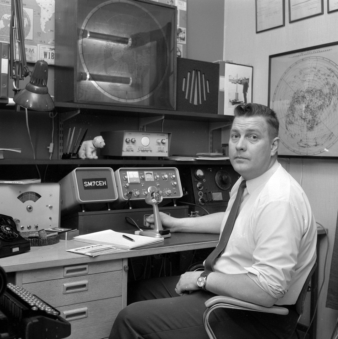 En man som sänder närradio sitter vid sitt skrivbord framför en mikrofon. Skrivbordet är fullt med radioutrustning. Han tittar in i kameran. Svartvitt foto.