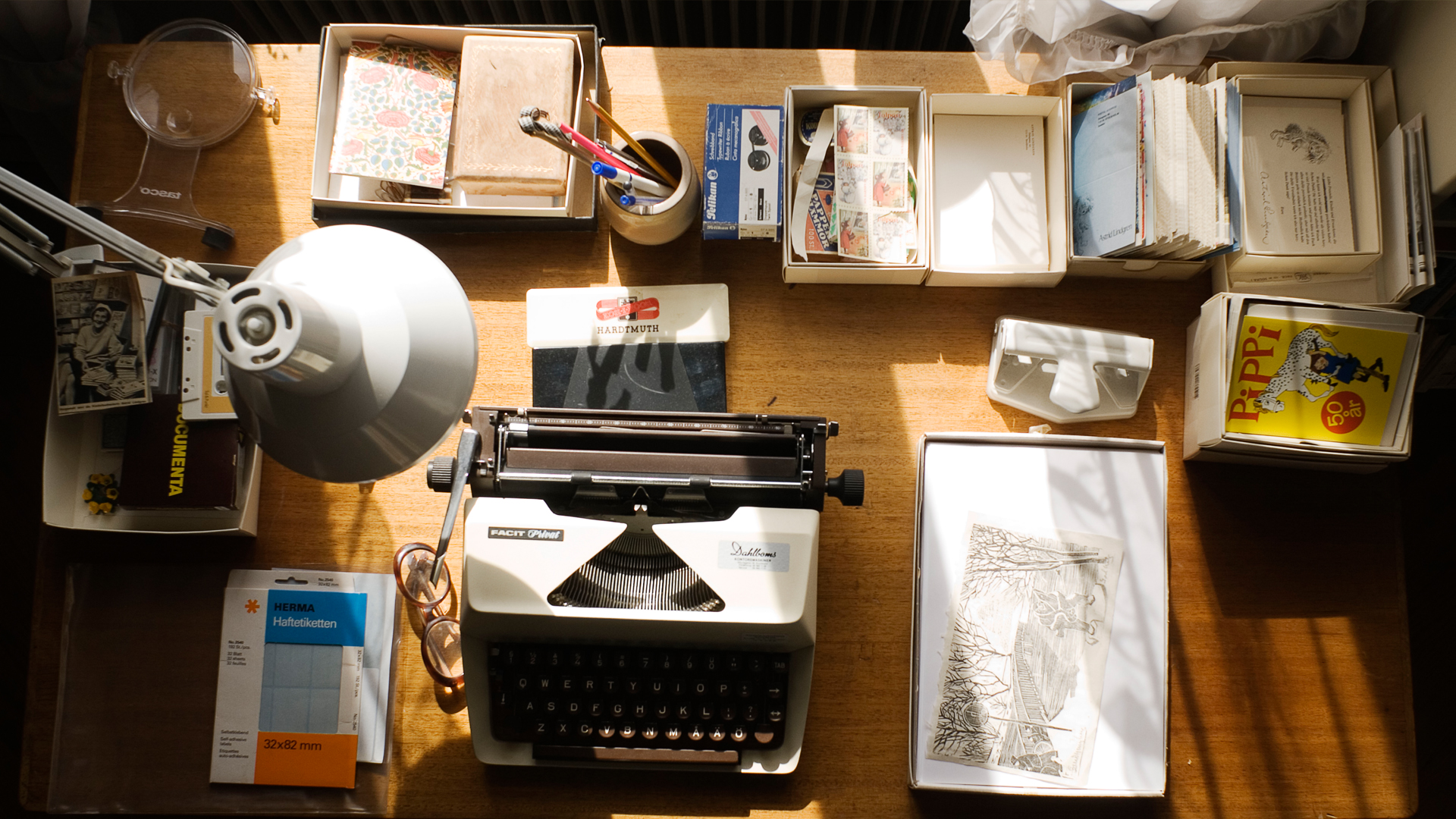 Ett skrivbord med olika saker. I mitten står en skrivmaskin och runt omkring finns små lådor med bland annat vykort och kassettband. Ett kort föreställer Pippi Långstrump och ett annat Astrid Lindgren.
