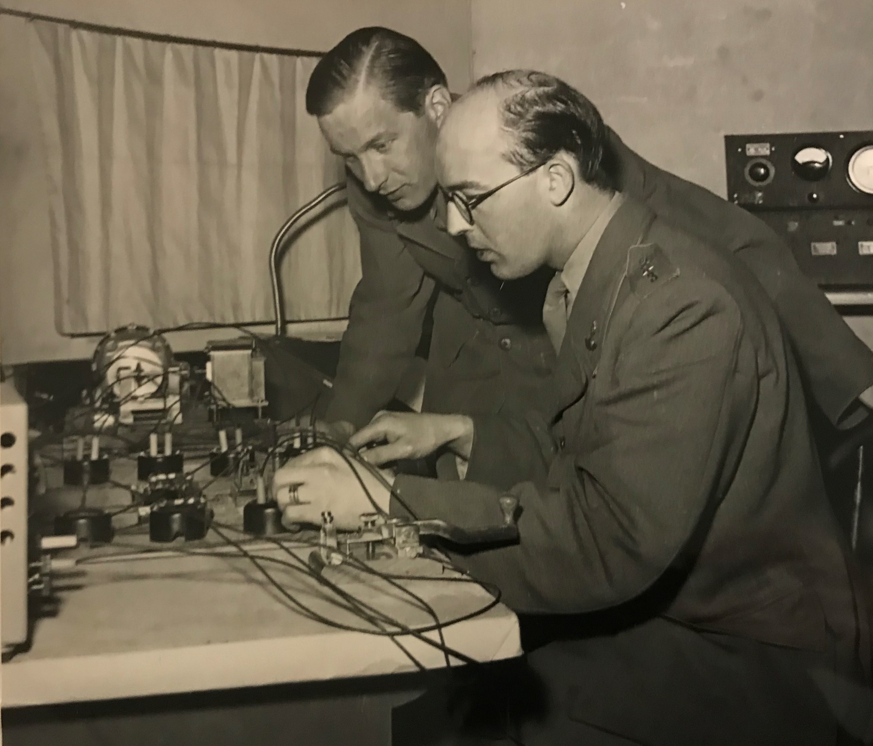 Två män iklädda militära uniformer står respektive sitter vid ett bord med radioutrustning.