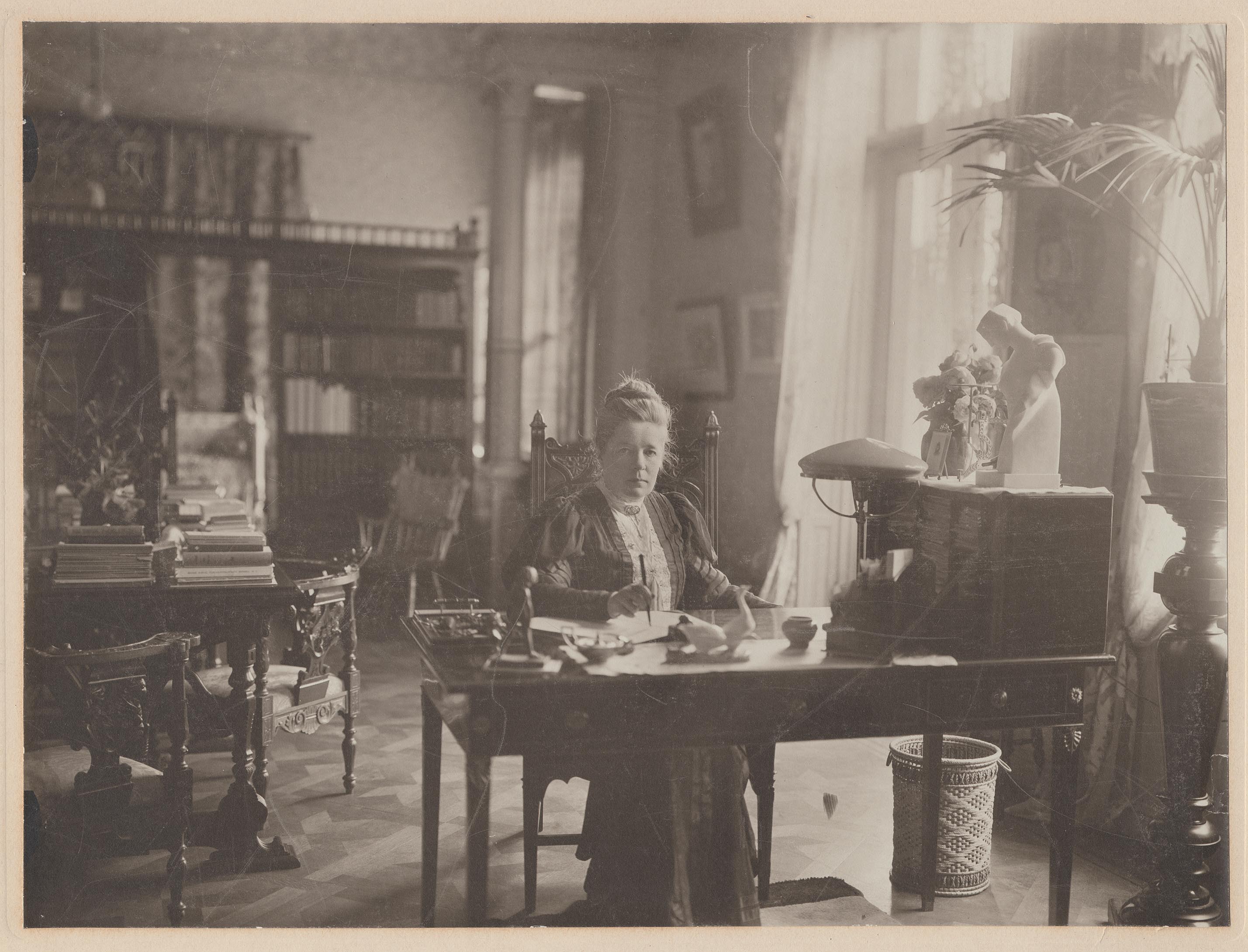 Selma Lagerlöf sitter vid ett skrivbord omgiven av snidade möbler, byster och krukväxter.