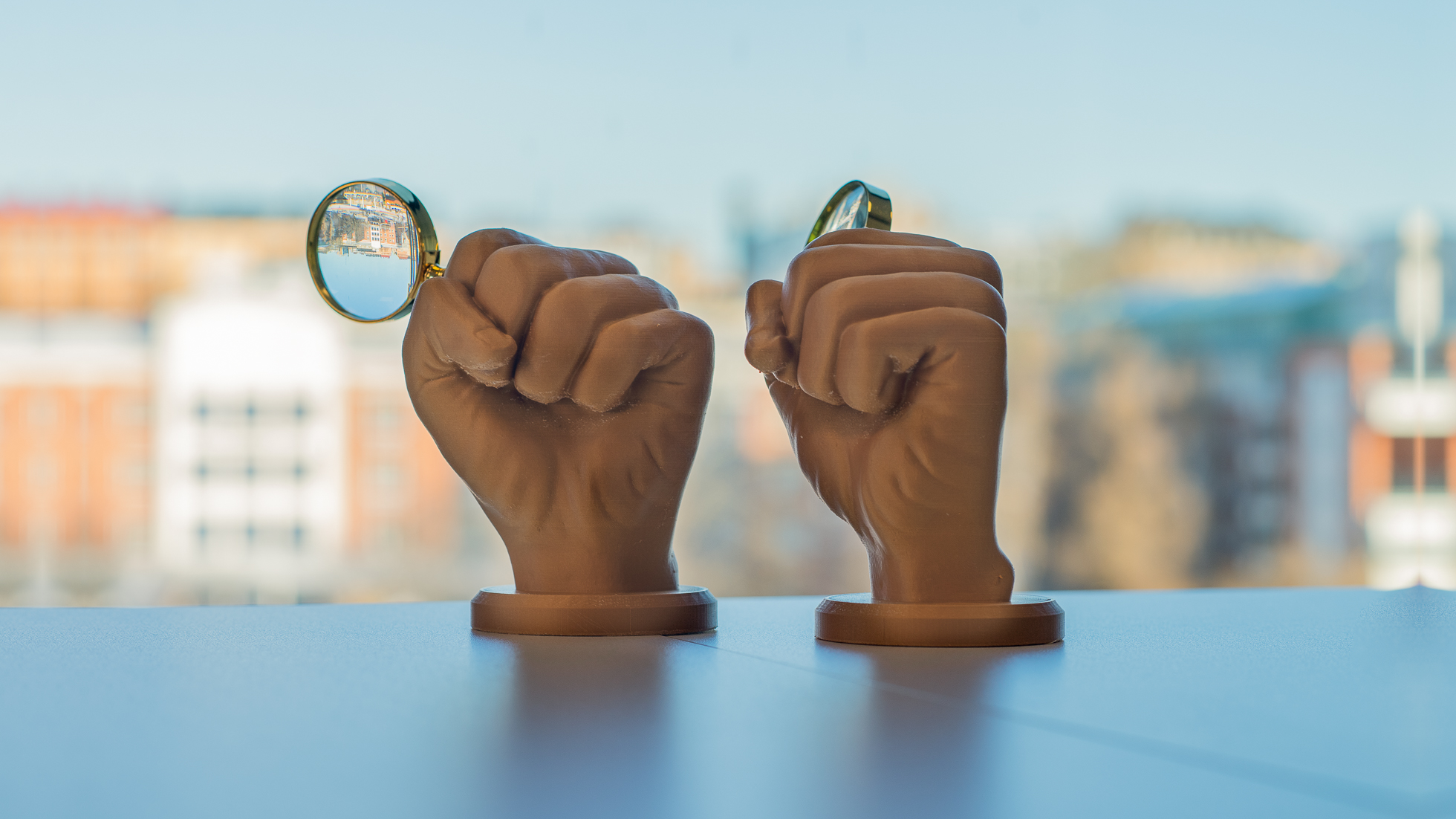 Två konstgjorda händer håller varsitt förstoringsglas mot bakgrund av ett fönster som visar en suddig vy över en stad.