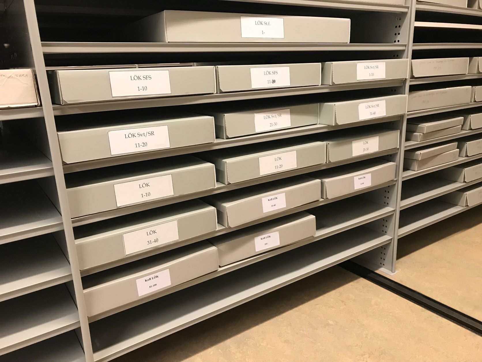Femton grå kapslar med vita etiketter innehållande julkalendrar, vilka är placerade på sex hyllplan i Kungliga bibliotekets underjordiska magasin